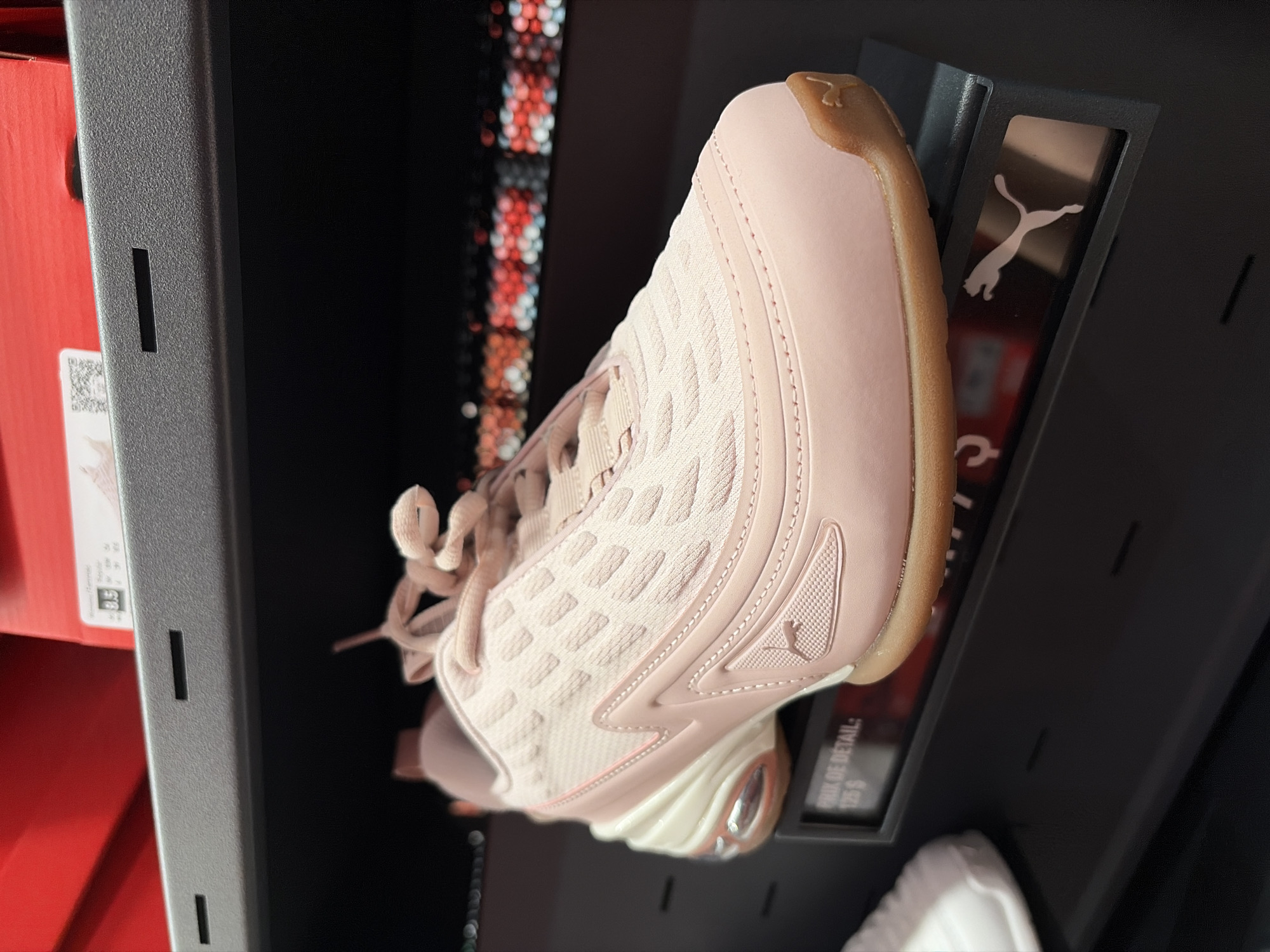 A light pink Puma athletic shoe is displayed on a black shelf in front of a blurred background of red boxes and lights. The shoe is positioned at an angle, showcasing its side profile. The shoe features a textured upper, a white and cream sole with a visible air bubble in the heel, and a gum-colored outsole. A small price tag with the text "PRIX DE DETAIL: 125 $" and a Puma logo are visible on the shelf beneath the shoe. The overall scene appears to be in a retail store environment.