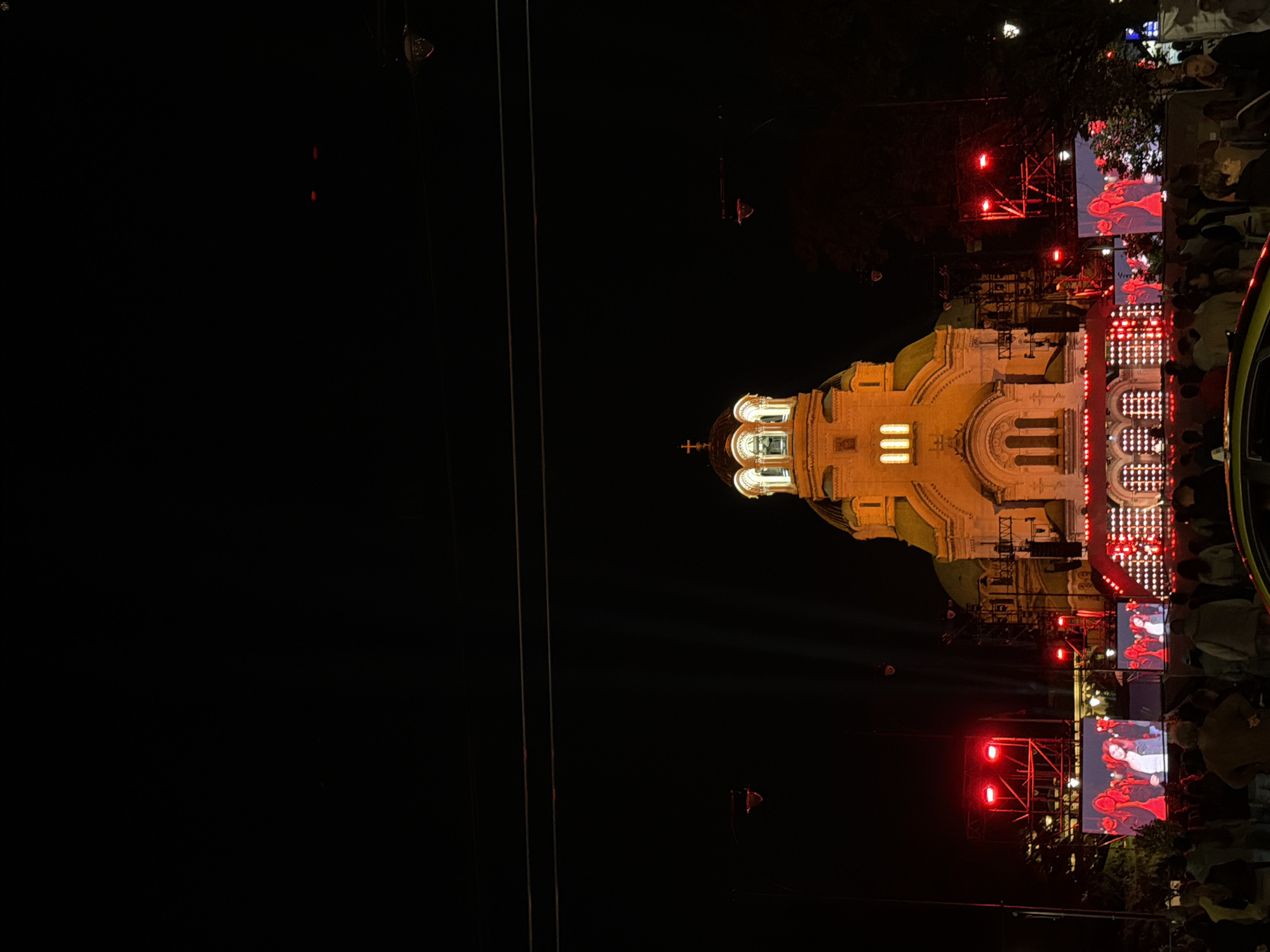 The image depicts a night scene in Sofia, Bulgaria, with the St. Alexander Nevsky Cathedral prominently illuminated and serving as a backdrop. In front of the cathedral, a large outdoor stage setup is visible, featuring multiple large LED screens displaying images of people, likely performers or attendees. The stage is adorned with numerous red lights. A crowd of people is gathered in the foreground, observing the event. The sky is dark, with visible overhead wires and streetlights. The overall atmosphere suggests an outdoor concert or public gathering.