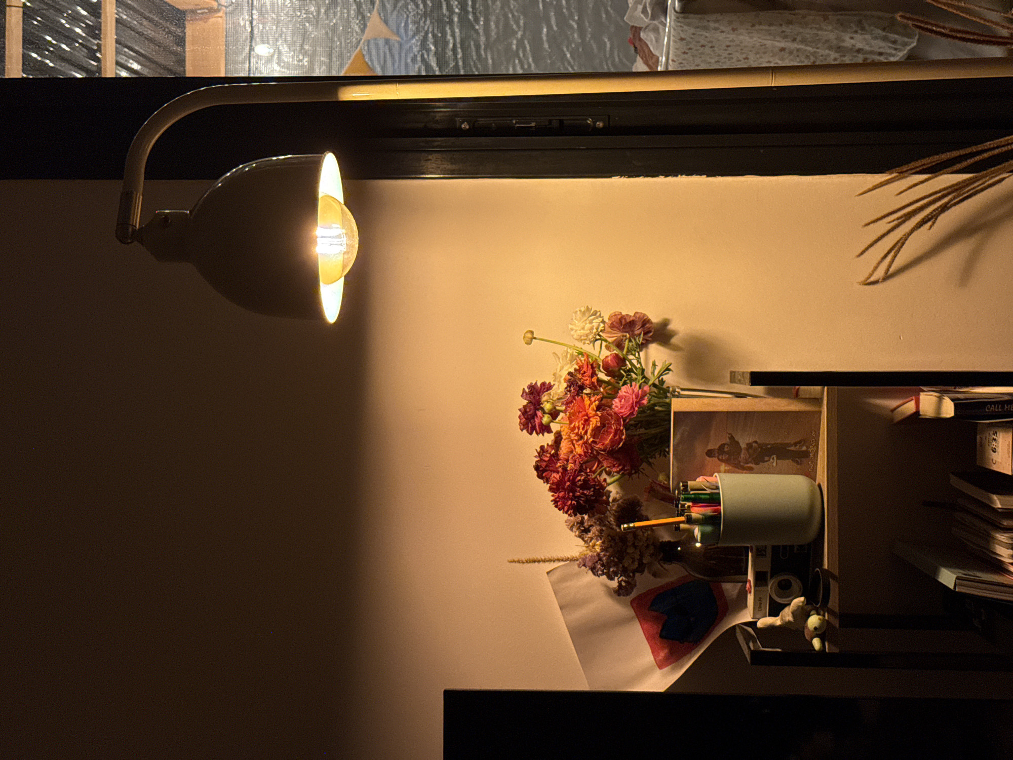 A close-up shot depicts a warm interior setting. A desk lamp is positioned to the left, casting light on the scene. A bouquet of flowers rests on a surface alongside a pencil cup filled with writing utensils. A framed picture is visible, and books are stacked on shelves. The location is not identifiable from the image, and there is no evidence to suggest it is in Tel-Aviv, Israel. No people are depicted in the scene.