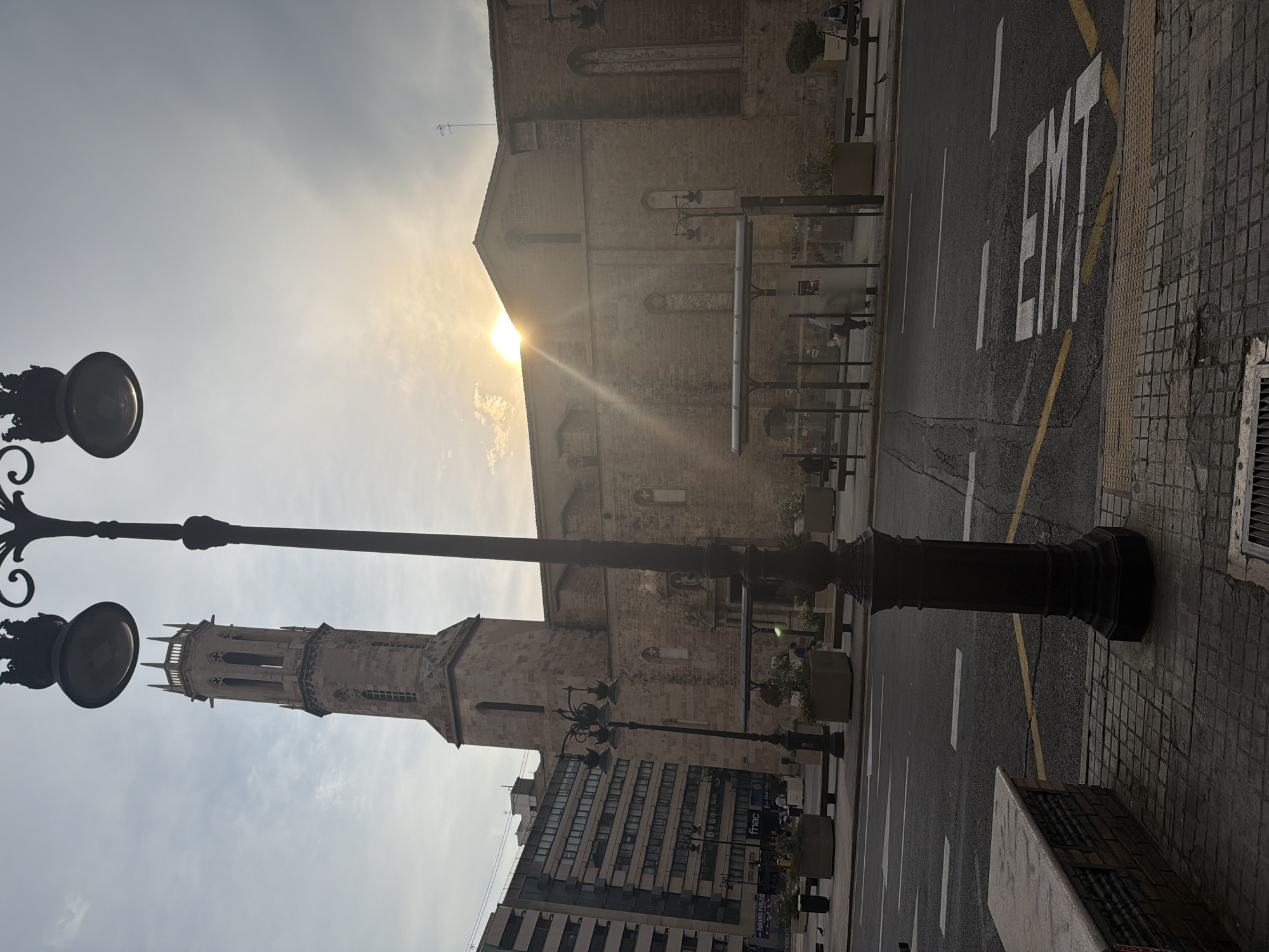 The image depicts a street scene in Valencia, Spain, featuring a prominent church tower and building with a stone facade. A vintage lamppost stands in the foreground, casting shadows. The sun is setting or rising, with rays of light filtering through the sky and illuminating the scene. A bus stop shelter is visible on the sidewalk, with a few people standing or walking nearby. The street is paved with asphalt and marked with white lines, indicating traffic lanes. The architecture suggests a historical part of the city.
