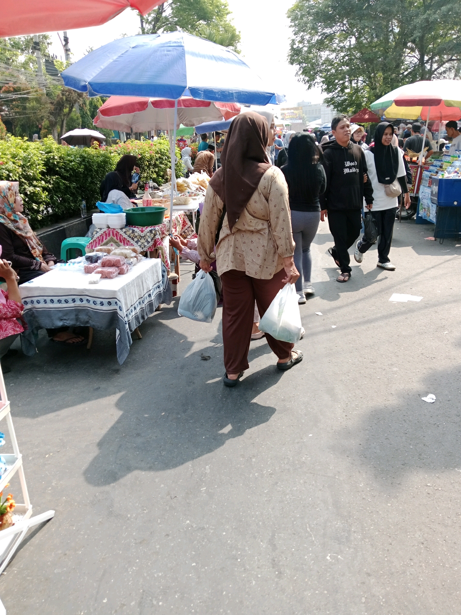 The image depicts a bustling outdoor market scene in Cianjur, Indonesia. Multiple individuals are visible, some browsing at stalls and others walking through the crowded pathway. A prominent foreground subject, a woman in a brown hijab and patterned long-sleeved top, walks away from the camera carrying a white plastic bag. She is wearing brown pants and dark sandals. To her left, another woman in a colorful hijab and pink top is partially visible, seated at a table laden with goods wrapped in plastic.
In the middle ground, several other people are seen walking. One man wears a black hoodie with "Wood Maiden" printed on it, and a woman beside him wears a white hijab and a black top. Stalls with umbrellas are set up, displaying various items, likely food and merchandise. One stall features a table covered with a blue and white patterned cloth, displaying an assortment of plastic-wrapped food items. Further in the background, more stalls and people are visible, suggesting a lively market environment. Trees and foliage line the background, and the ground is paved asphalt. The sun is casting shadows, indicating it is daytime.