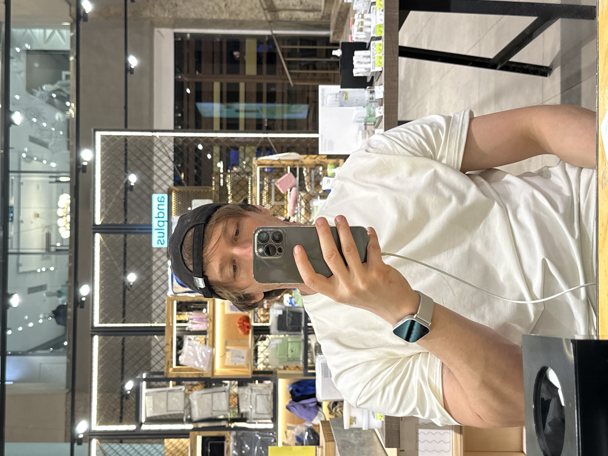 A man is taking a selfie in front of a mirror. He is wearing a white t-shirt and a black baseball cap with the word "GULLIVER" printed on it. He is holding a phone in his hand, and there is a charging cable plugged into it. He is wearing a smartwatch.
The reflection in the mirror shows various displays of merchandise in a retail setting. There are shelves containing various products, including handbags and other accessories. The lighting is bright, and the overall scene appears to be a commercial environment. The Gulliver sign on the cap suggests the location may be a Gulliver store, a popular chain in Moscow, Russia.