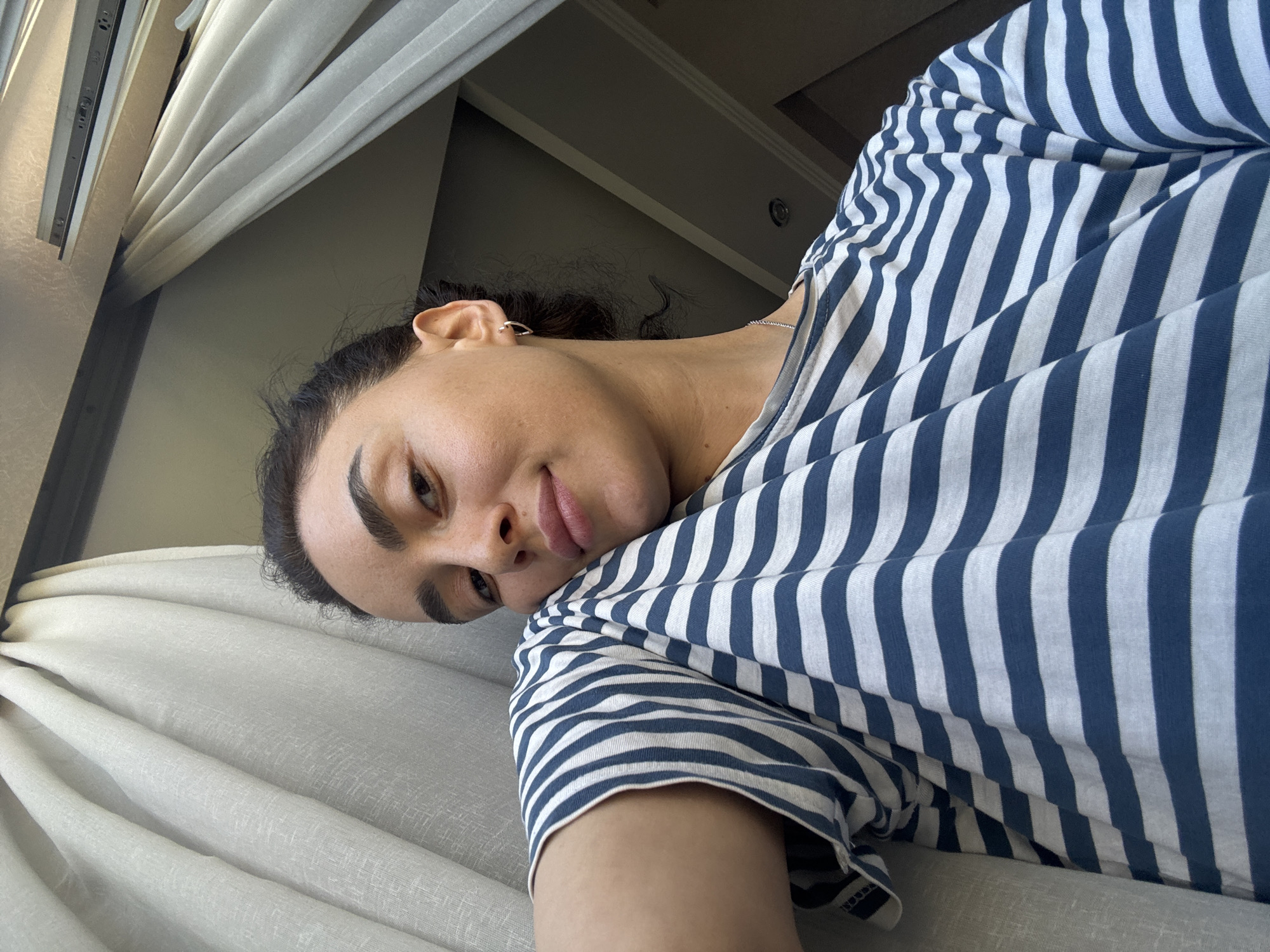 A woman with dark hair pulled back and prominent eyebrows is leaning on her arm, looking towards the camera with a soft smile. She is wearing a blue and white striped t-shirt and a thin silver necklace. The scene appears to be indoors, with sheer white curtains to her left and a glimpse of a window frame and ceiling above. The lighting suggests it is daytime. The specific location within the city of Tbilisi, Georgia, is not identifiable from the visual information provided.