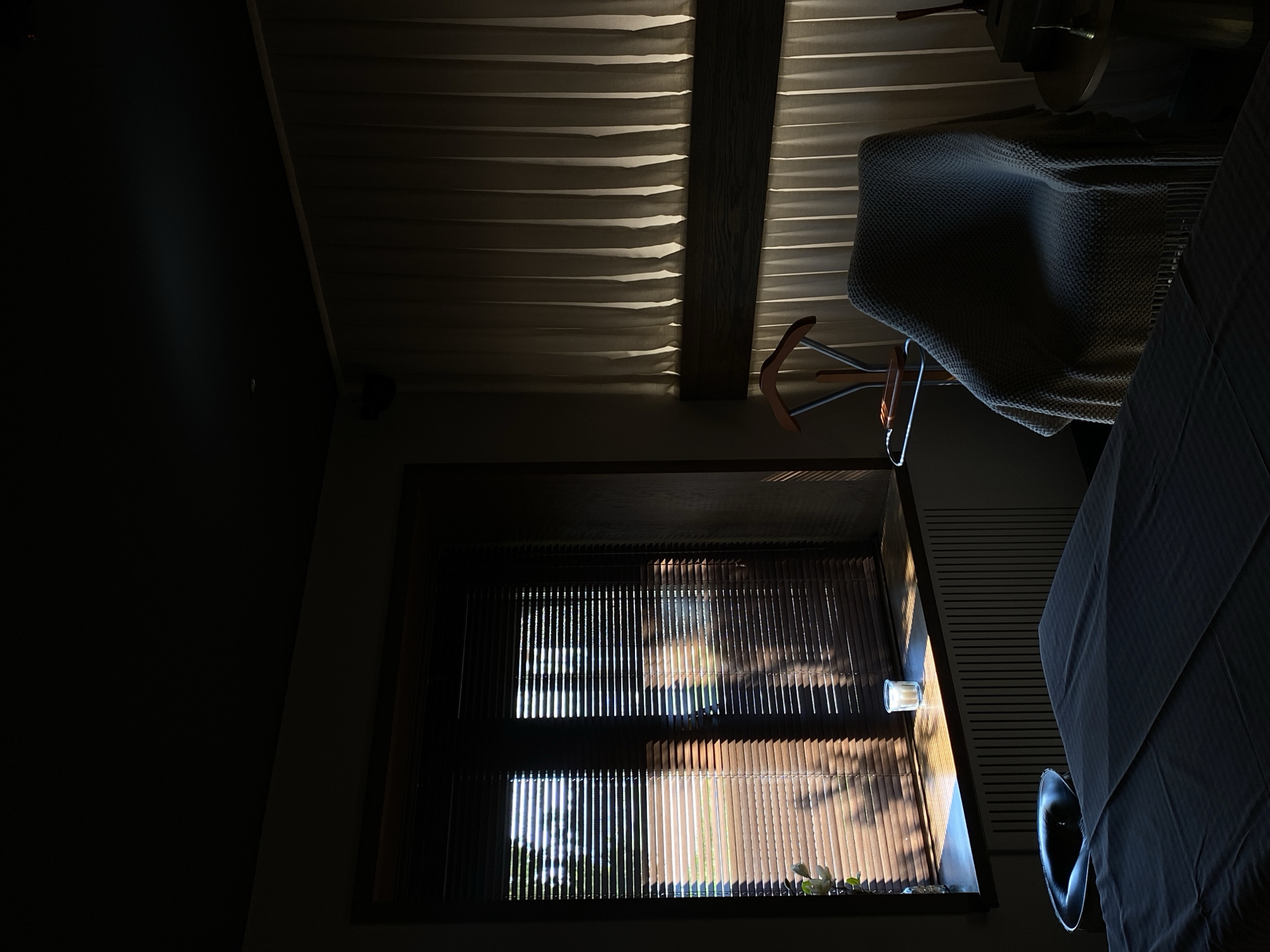 The image shows an interior scene, likely taken in Votkinsk, Russia (though there is no evidence to confirm this). The room is dimly lit, with sunlight streaming through a window covered by blinds. The light creates stark contrasts of light and shadow across the surfaces. Visible within the room are a wooden beam structure on the ceiling, a chair with a knitted blanket draped over it, and a table covered with a tablecloth. On the windowsill are a few plants, partially illuminated by the sun. The left side of the frame is dominated by darkness.