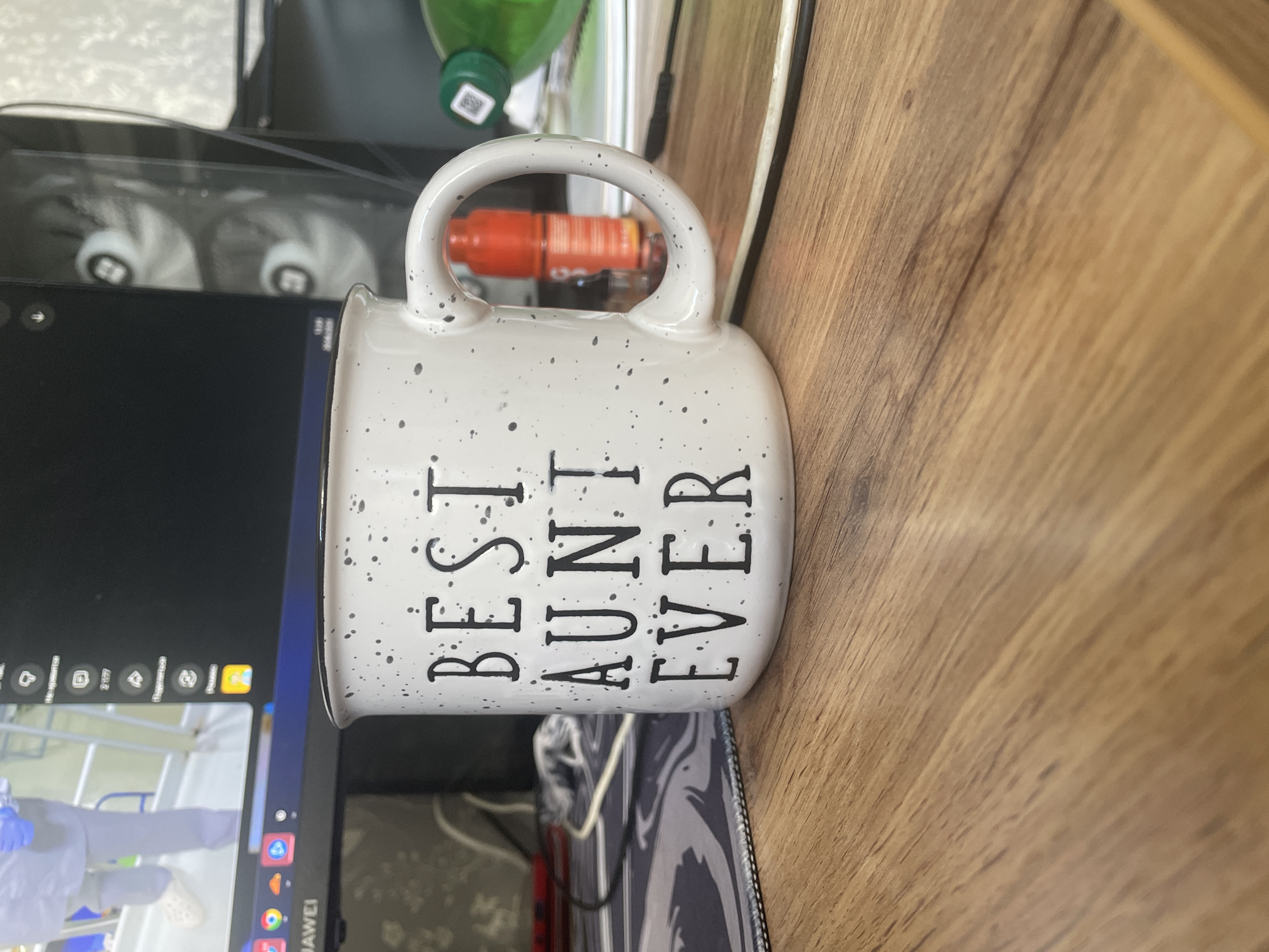 A speckled ceramic mug with the words "BEST AUNT EVER" in black lettering sits on a wooden desk. The mug is positioned in the foreground, slightly to the left of center. Behind the mug, a computer monitor is visible, displaying various icons and what appears to be a video feed. To the right of the mug, there's a red bottle and a green bottle with a white label. The desk surface is made of light brown wood with a visible grain pattern. The background includes a computer tower with visible cooling fans and wires. There are no people clearly depicted in the scene, though a blurry figure is visible on the computer screen. The overall setting suggests an indoor workspace. The location is identified as Sinyavino, Russia.
