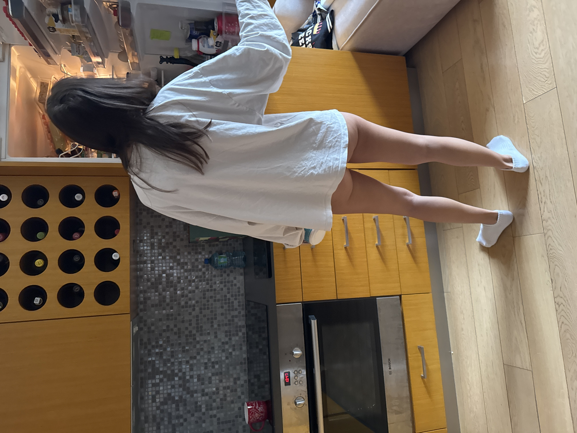 A woman with dark hair is standing in a kitchen in what appears to be an apartment or home. She is wearing a white oversized shirt and white socks, and is reaching into an open refrigerator. The kitchen has wooden cabinets, a gray tiled backsplash, a wine rack, and a built-in oven. The floor is wooden. The time displayed on the oven is 13:47. Based on architectural styles and general appearance, this kitchen may be located in a modern apartment in a city like Warsaw, Poland.