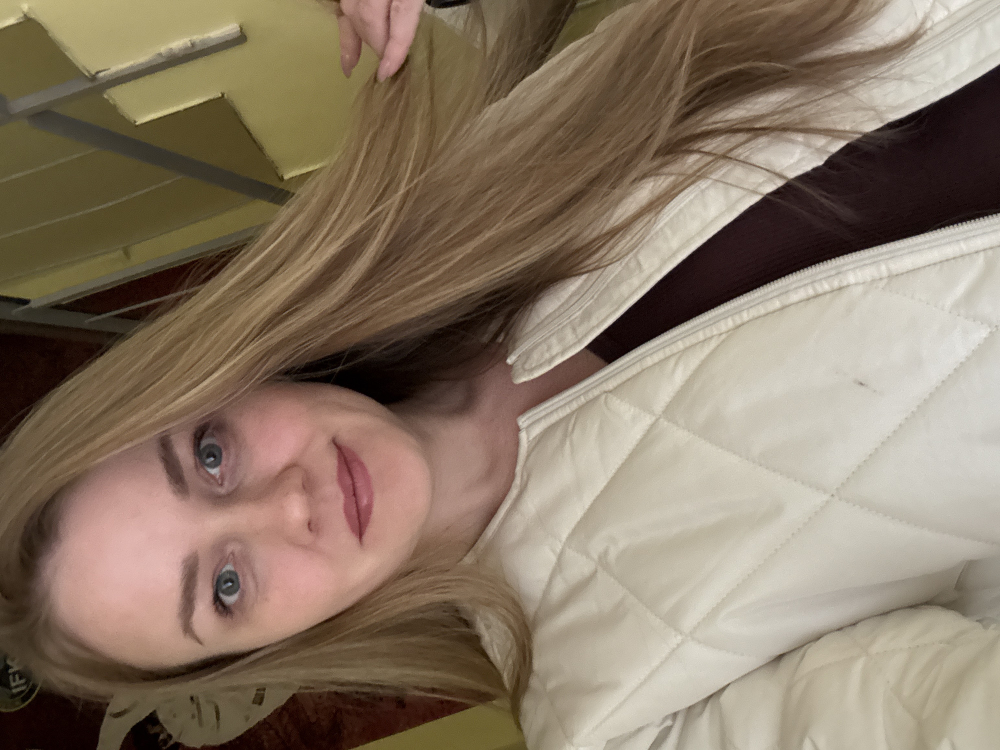 The image shows a woman with long, blonde hair and blue eyes wearing a white, quilted jacket over a dark, possibly maroon-colored top. She is looking directly at the camera with a slight smile. The background is blurry and out of focus, but reveals parts of a ceiling and other objects suggesting an indoor setting. The exact location is not discernible from the image and therefore its location in Chaikovsky, Russia, cannot be confirmed.
