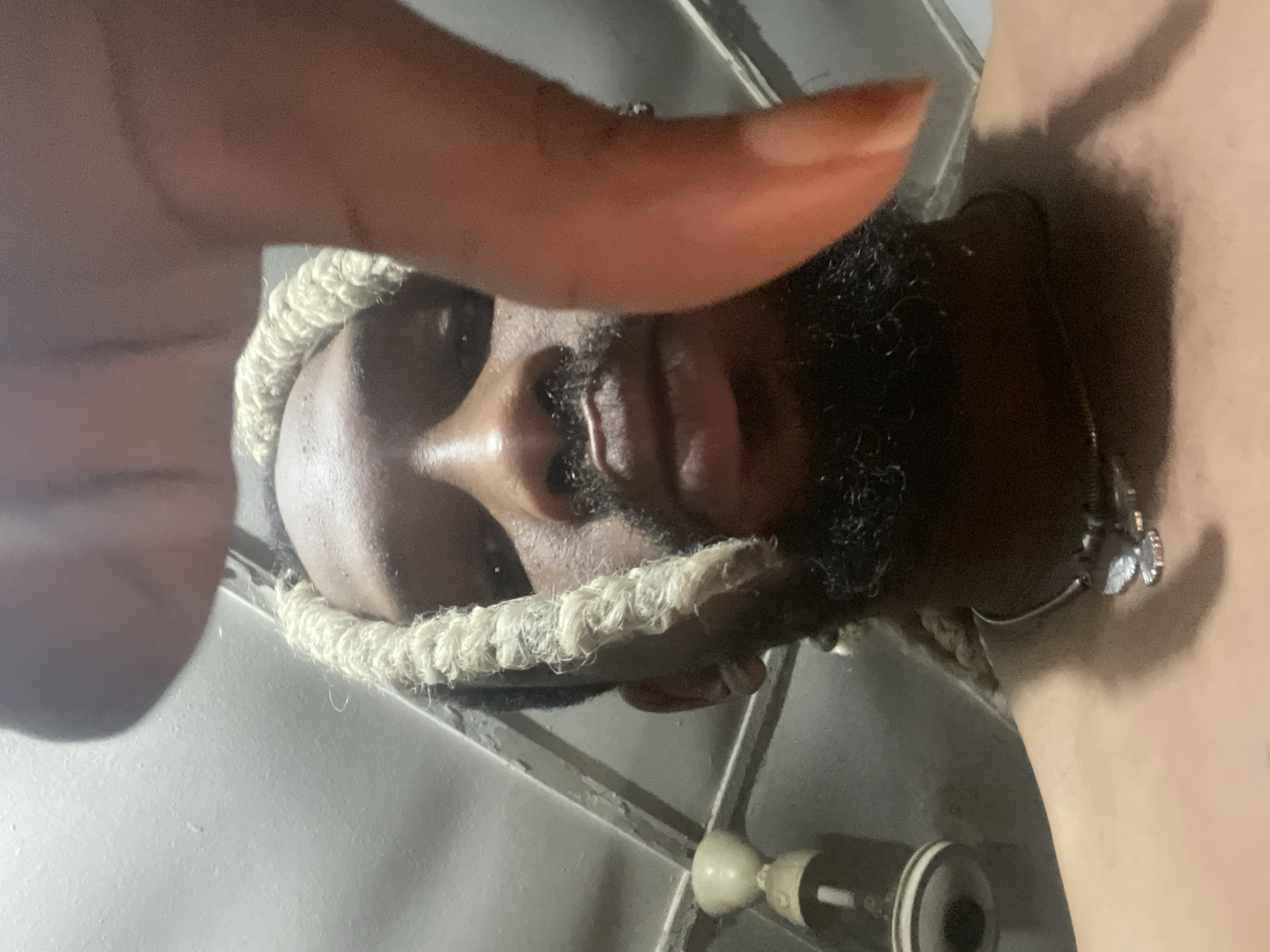 The image shows a close-up, low-angle, selfie-style shot of a Black male subject with blonde dreadlocks and a beard. The subject is looking downwards towards the camera, with a thumb obscuring part of the frame from the top right. The subject is wearing a necklace with a silver pendant. The background is dimly lit and appears to be an indoor environment, possibly a room with a ceiling fan visible on the left. The overall impression is of a casual and personal moment. However, there is no specific visual information in the image that can be used to determine the location as Jos, Nigeria.