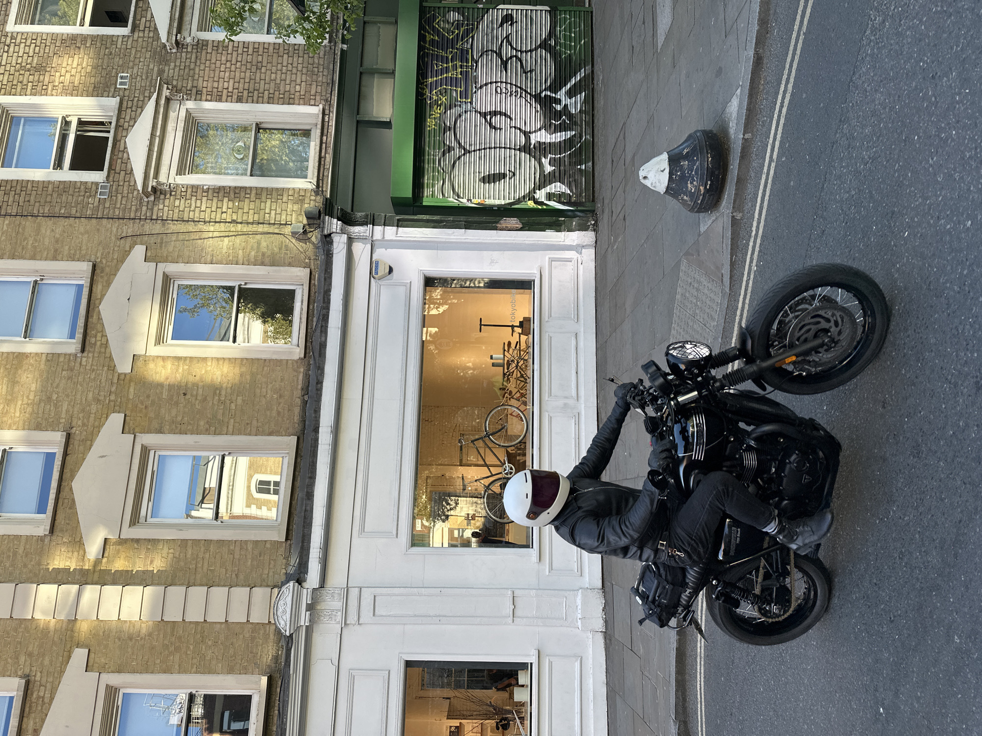 An overhead, slightly angled view captures a street scene in London, United Kingdom, on a sunny day. A person wearing a black leather jacket, black trousers, black boots, and a white and maroon full-face helmet is seated on a black motorcycle, appearing stationary on the asphalt road next to the curb. A black backpack is visible on the motorcycle's rear. The motorcycle has spoked wheels and visible disc brakes.
To the left, a traditional brick building with multiple windows dominates the scene. The building features ornate window sills and decorative cornices between floors. Reflections of trees and blue sky are visible in the windowpanes. Below this, a white shopfront with large display windows occupies the ground level. Inside the shop window, a bicycle is prominently displayed, and faint text reading "TOKYOBIKE" is visible on the glass, suggesting a bicycle shop. Above the shop, a closed green corrugated metal gate is covered in white and black graffiti.
The road features double white lines, and a distinctive black and white bollard is positioned on the sidewalk near the edge of the road. The street and sidewalk appear clean and well-maintained. Strong sunlight casts clear shadows, indicating a bright daytime setting.