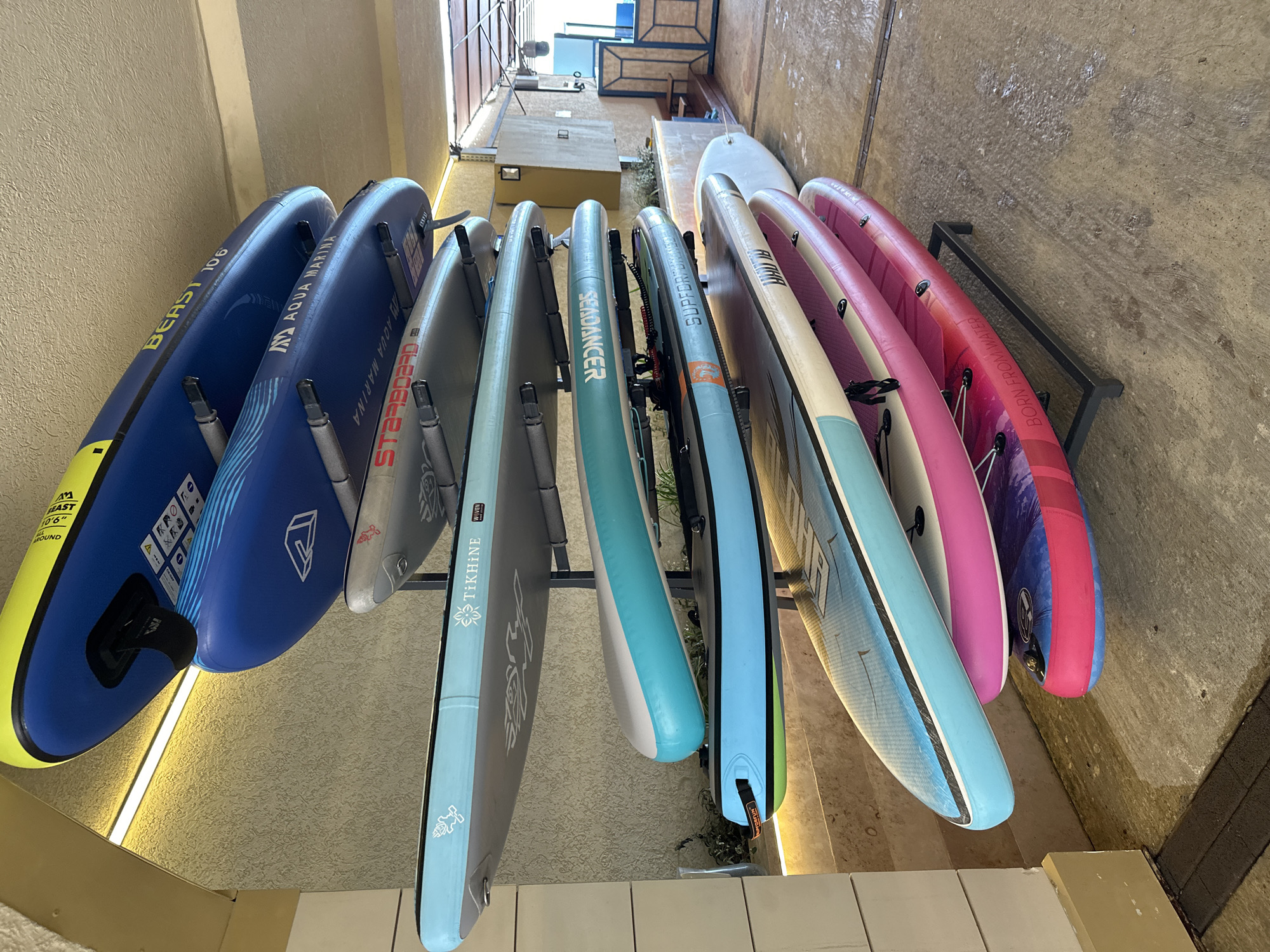 A rack of stand-up paddleboards is stored outdoors under a covered area with concrete flooring. The paddleboards are stacked vertically, with each board resting on metal supports. The boards have various colors and designs, including blue, turquoise, white, pink, and purple. Some boards have brand names and logos visible, such as "BEAST 106," "AQUA MARINA," "TiKHINE," "WOVEN," and "SEA DANCER." The environment appears to be a storage facility or a beachside rental area. The lighting suggests it is daytime, with artificial lights visible on the ceiling. In the background, there is a building wall with textured plaster, some greenery, and part of a structure with metal fencing and wooden panels. The location is identified as Sochi, Russia.