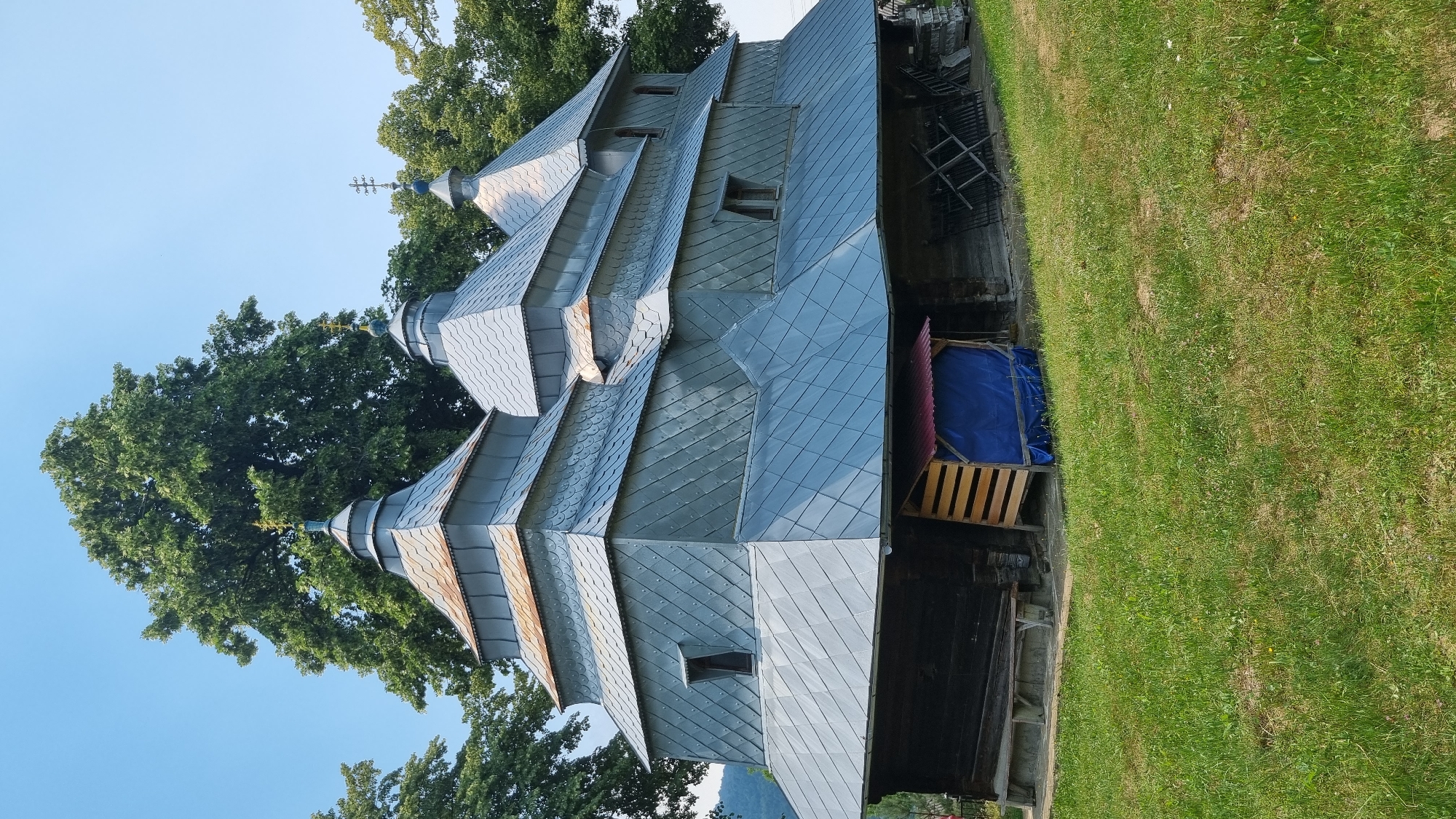 The image depicts a wooden church with multiple tiered roofs covered in grey metal shingles. The church has a dark wooden lower section. In front of the church, there is a simple wooden bench on the left and a small wooden structure with a red roof covered by a blue tarp on the right. The ground in front of the church is covered in green grass. Behind the church, there are tall green trees and a clear blue sky. The overall scene suggests a rural setting, likely in Vyshka, Ukraine, given the architectural style often found in that region. There are no people visible in the image.