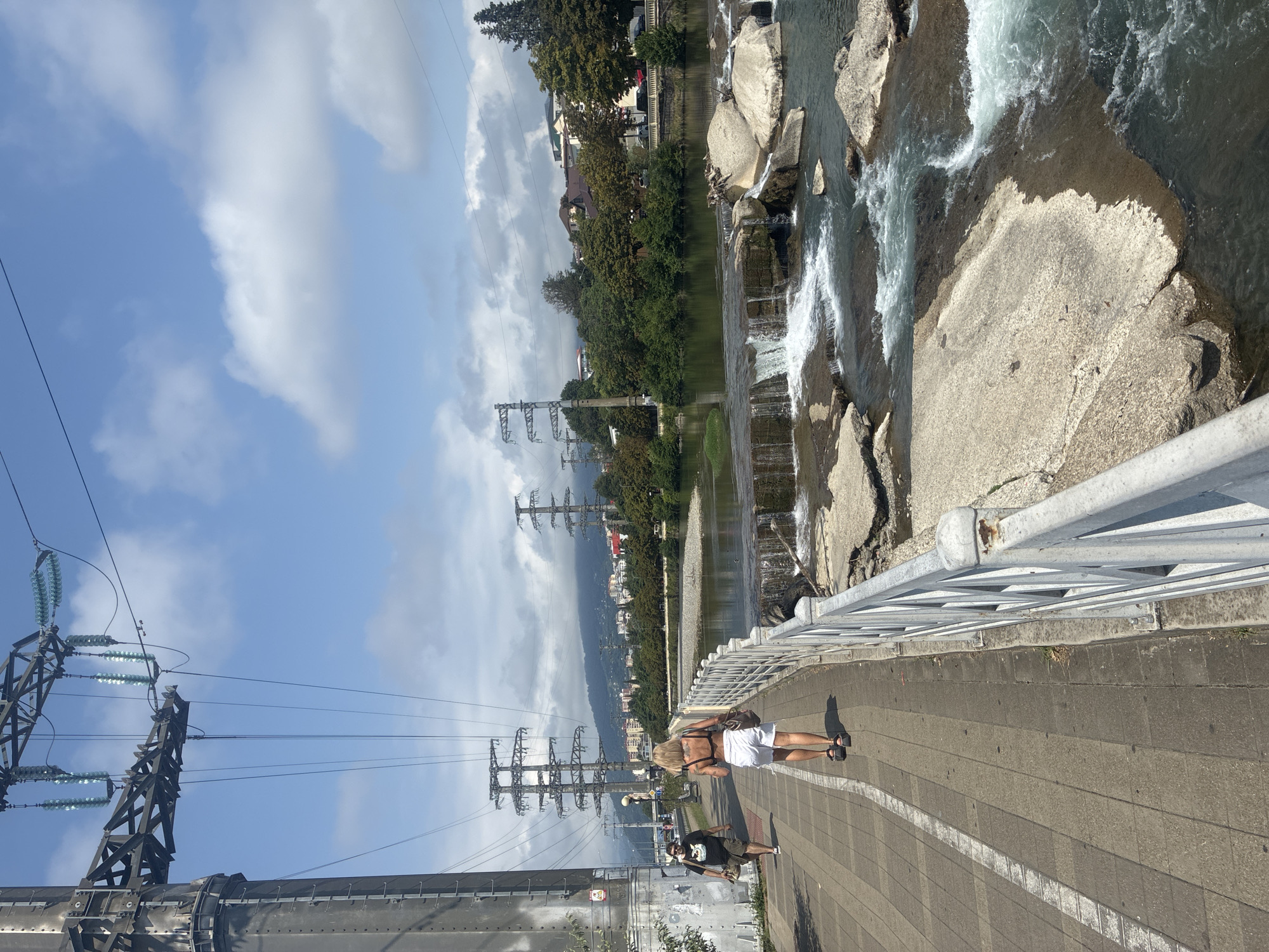 An outdoor, eye-level, full shot captures a scenic view along a river in Sochi, Russia, with a promenade in the foreground. On the left side of the frame, a large, grey electrical tower looms, adorned with insulators and power lines stretching across the sky. Two individuals are walking away from the camera on the promenade: a woman with blonde hair wearing a black bikini top and white shorts, and a man in a black t-shirt and olive-green shorts. A white metal railing separates the promenade from the river, which features a small waterfall cascading over rocks and an array of light grey, tan, and brown stones. The riverbank is lined with lush green trees and buildings in the background, with more electrical towers and a hazy mountain range on the horizon under a partly cloudy sky.