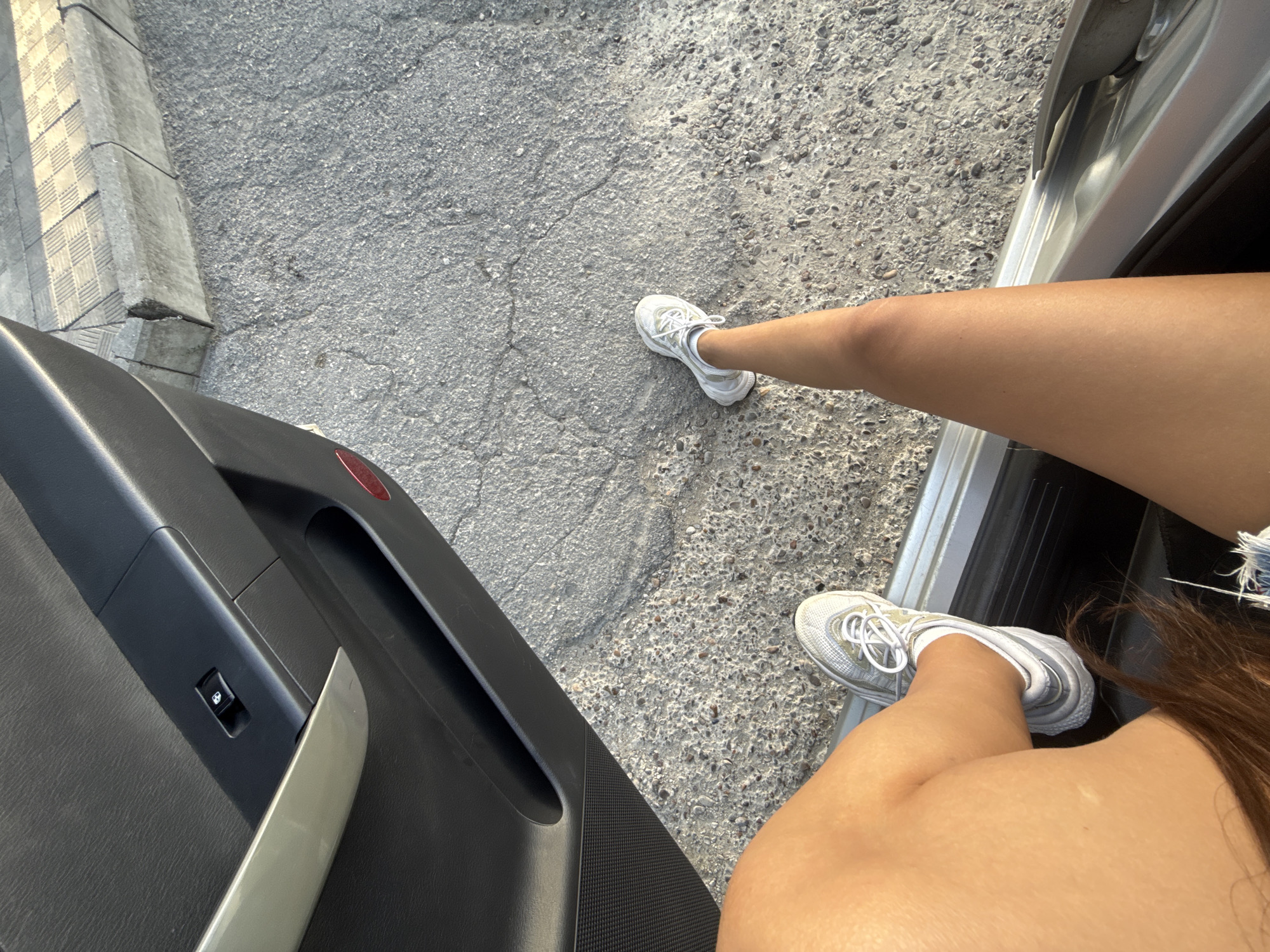 The image shows a person's legs and feet from a high angle, sitting in the open doorway of a car. The person is wearing white athletic shoes with gray accents and white laces. The left leg is bent, with the foot resting on the ground. The right leg is extended, with the foot also on the ground. The car interior is visible, including the black door panel with a silver handle and a window control button. The exterior environment appears to be a paved road or sidewalk with some debris and a concrete curb with textured paving stones behind it. The lighting suggests it is daytime, possibly late afternoon due to the angle of the shadows. There are no visible landmarks that definitively place this scene in Cimișlia, Moldova.