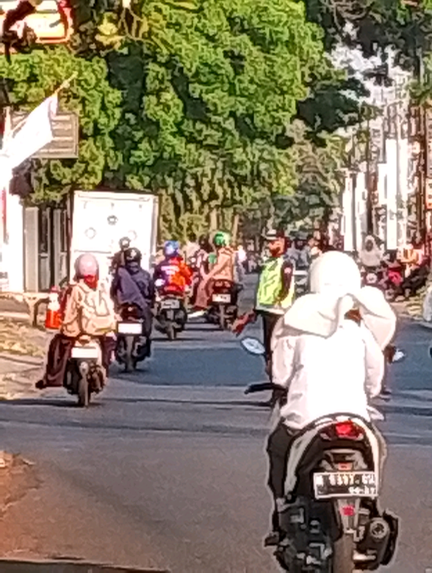 The image depicts a street scene in Probolinggo, Indonesia, characterized by a procession of motorcyclists. The foreground is dominated by the rear view of a motorcyclist wearing a white helmet and a white long-sleeved shirt. This motorcycle has a license plate that reads "B 3397 GZL".
The street is paved and appears to be an urban road, lined with buildings and lush green trees. Several other motorcyclists are visible in the middle ground, also traveling in the same direction. These include individuals wearing helmets of various colors such as pink, blue, and green, and others in casual attire or what appears to be a yellow safety vest over a darker outfit, suggesting they might be part of a local service or authority.
The motorcycles themselves are predominantly scooters, a common mode of transport in the region. The environment suggests a sunny day, with the sunlight casting shadows and highlighting the greenery. The overall impression is of a busy street, likely during daytime hours, with people commuting or traveling on motorcycles. The buildings on the side of the road appear to be commercial or residential structures, typical of an Indonesian city.