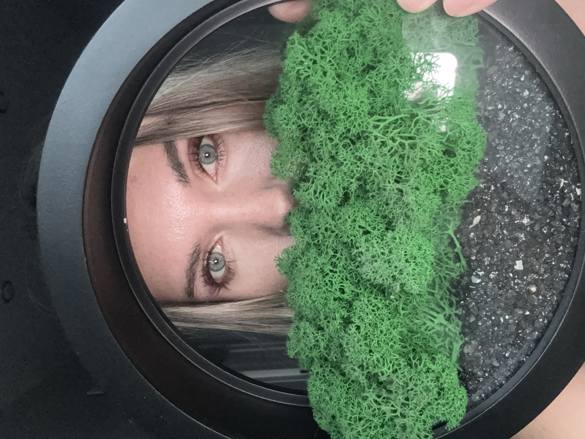 The image displays a close-up, rotated view of a person's reflection within a circular, framed object. The reflection prominently features the person's light blue-grey eyes, part of their nose, and a portion of their forehead and light-colored (blonde or ash blonde) hair. The expression appears calm and focused.
The circular object itself is encased in a dark, possibly black, frame. Inside, it contains two distinct materials. A significant portion is filled with a vibrant green, highly textured material that resembles preserved moss or lichen, exhibiting a dense, intricate, and branched structure. Adjacent to this green material, the remaining section of the circular object is filled with dark, granular material, consistent with small gravel or decorative stones, which includes some lighter, possibly reflective, particles.
A hand or fingers are visible at the top of the frame, suggesting the circular object is being held or adjusted. The scene implies an interaction with a decorative item that combines natural elements with a reflective surface, possibly a small terrarium or framed botanical art. No specific time of day, weather conditions, or explicit location cues are visible within the image, other than the provided context of Votkinsk, Russia. There is no visible text.