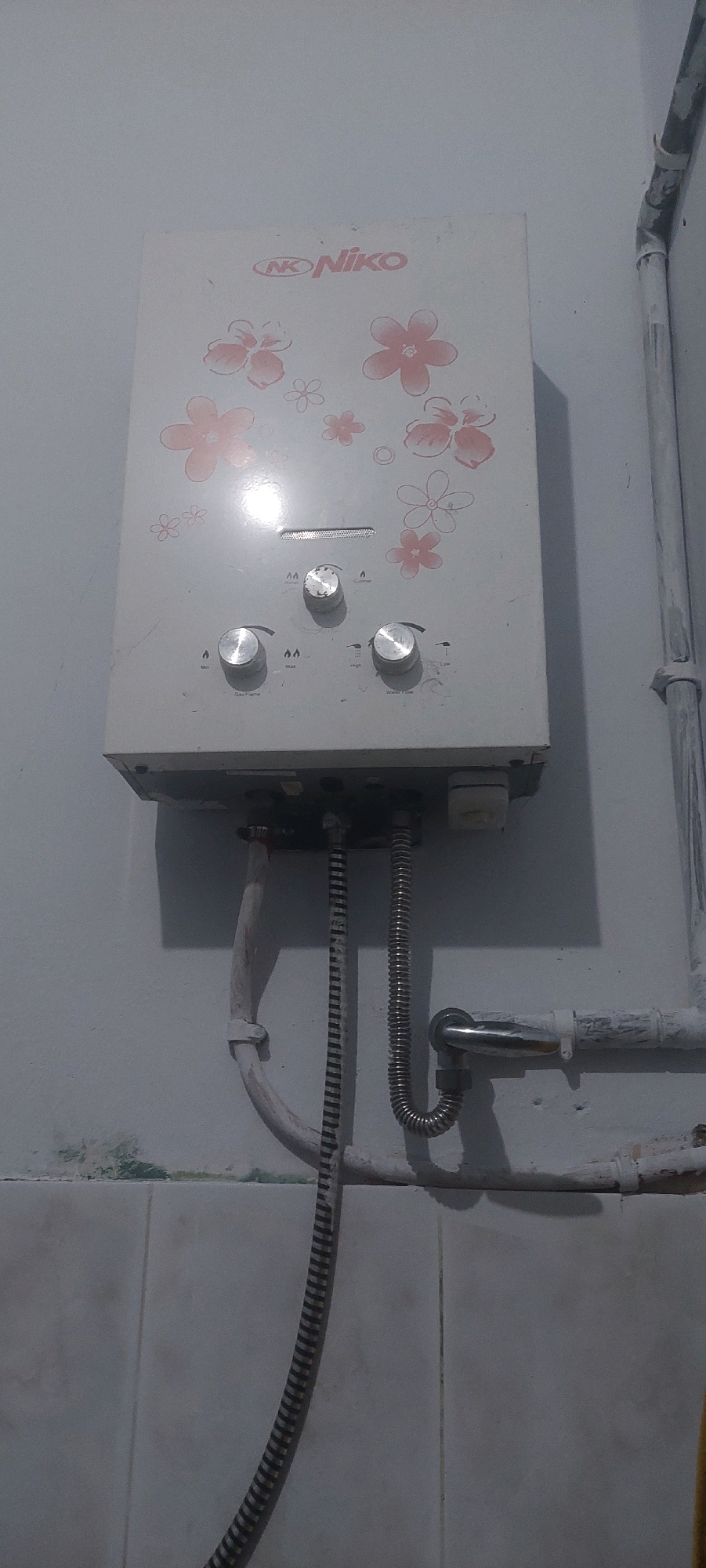 The image shows a gas water heater mounted on a wall. The water heater is white with pink floral patterns and has three knobs for control. It is connected to plumbing pipes. The wall appears to be painted white, with some tiles visible below. There are also some pipes running vertically and horizontally. The scene is likely an indoor bathroom or kitchen area. The image does not provide any specific clues about the location within Lumajang, Indonesia.