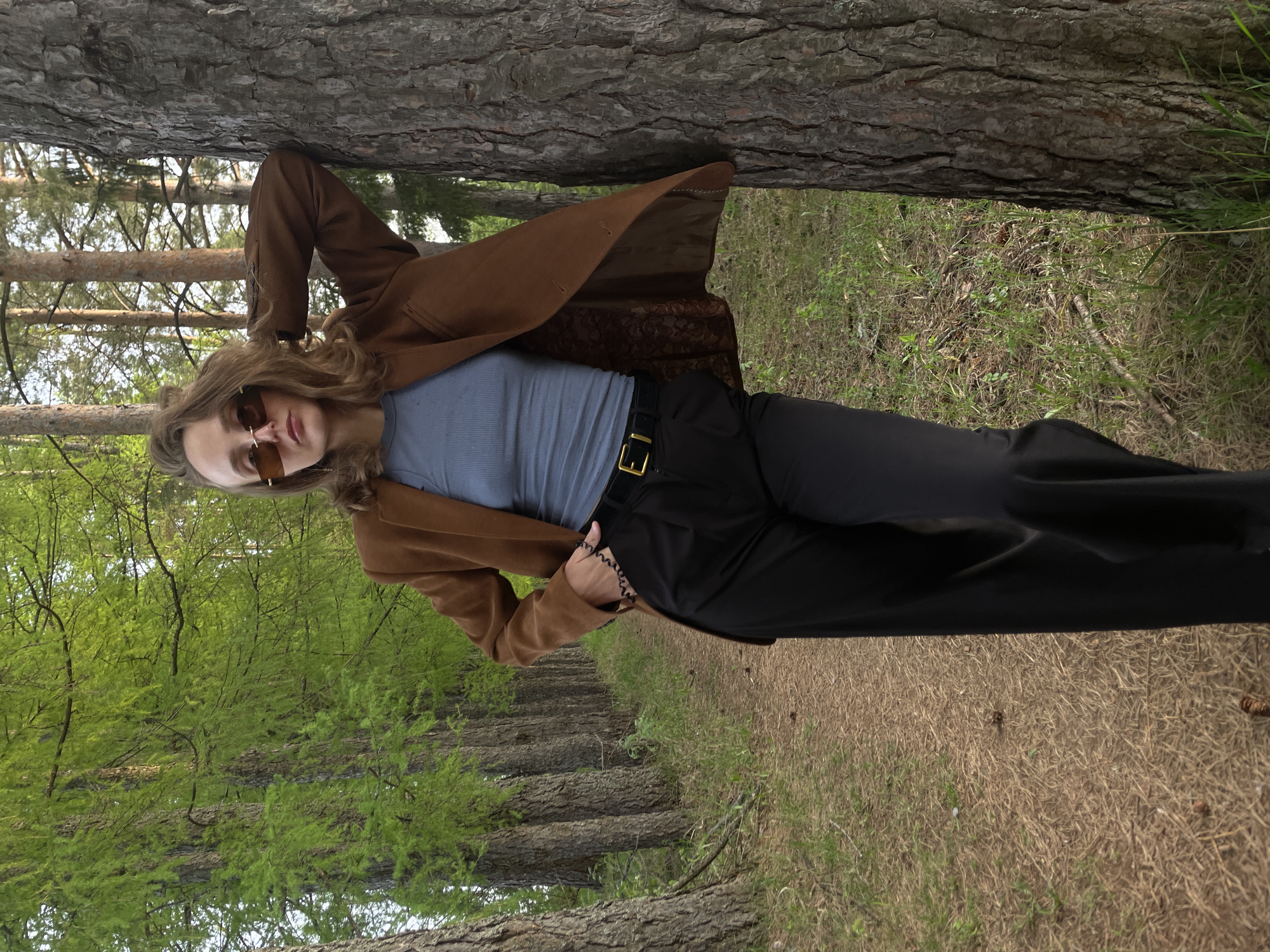 A woman in sunglasses, a brown coat, a blue shirt, and black pants is standing next to a tree in a forest or park in Подшивалово, Russia. The location is evident from the Russian place name. The ground is covered in grass and leaves. The trees in the background are spaced evenly. The woman is leaning against the tree with her right hand in her pocket. Her left arm is bent at the elbow and raised. The angle of the photograph suggests that it was taken from a low perspective.