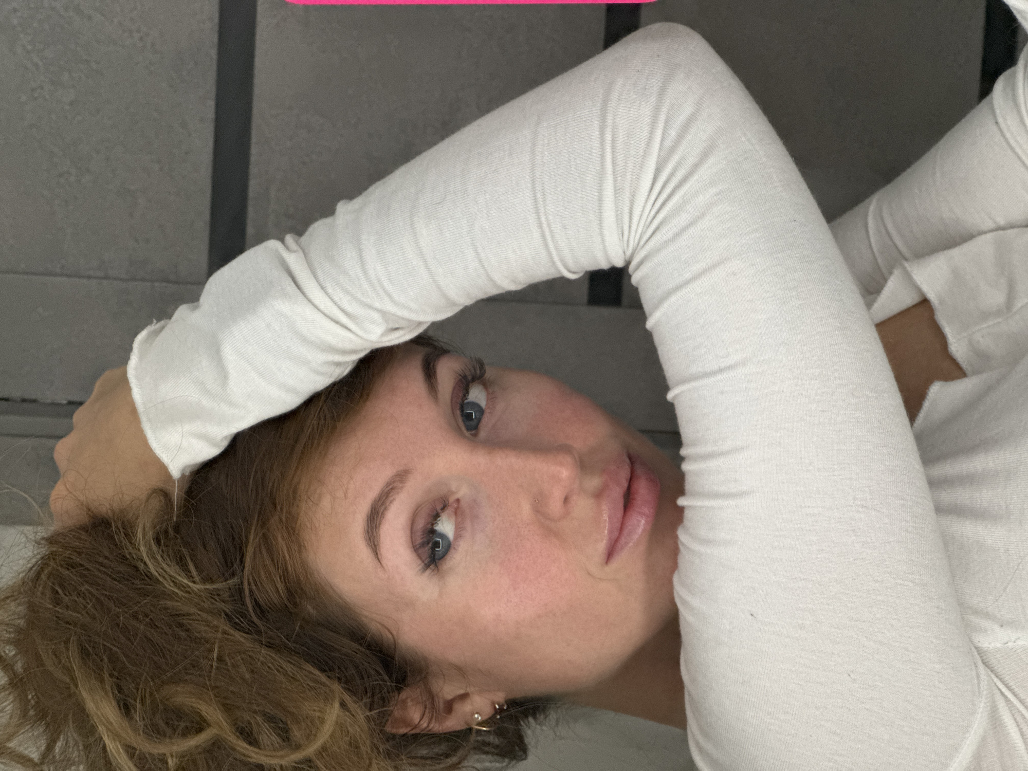 A young woman with light skin and blue eyes is lying down. She has curly brown hair. She is wearing a white, long-sleeved shirt. Her left arm is bent and resting near her head. The background is a gray wall with vertical lines. There is no indication that the location is in the city of Chaikovsky, Russia.