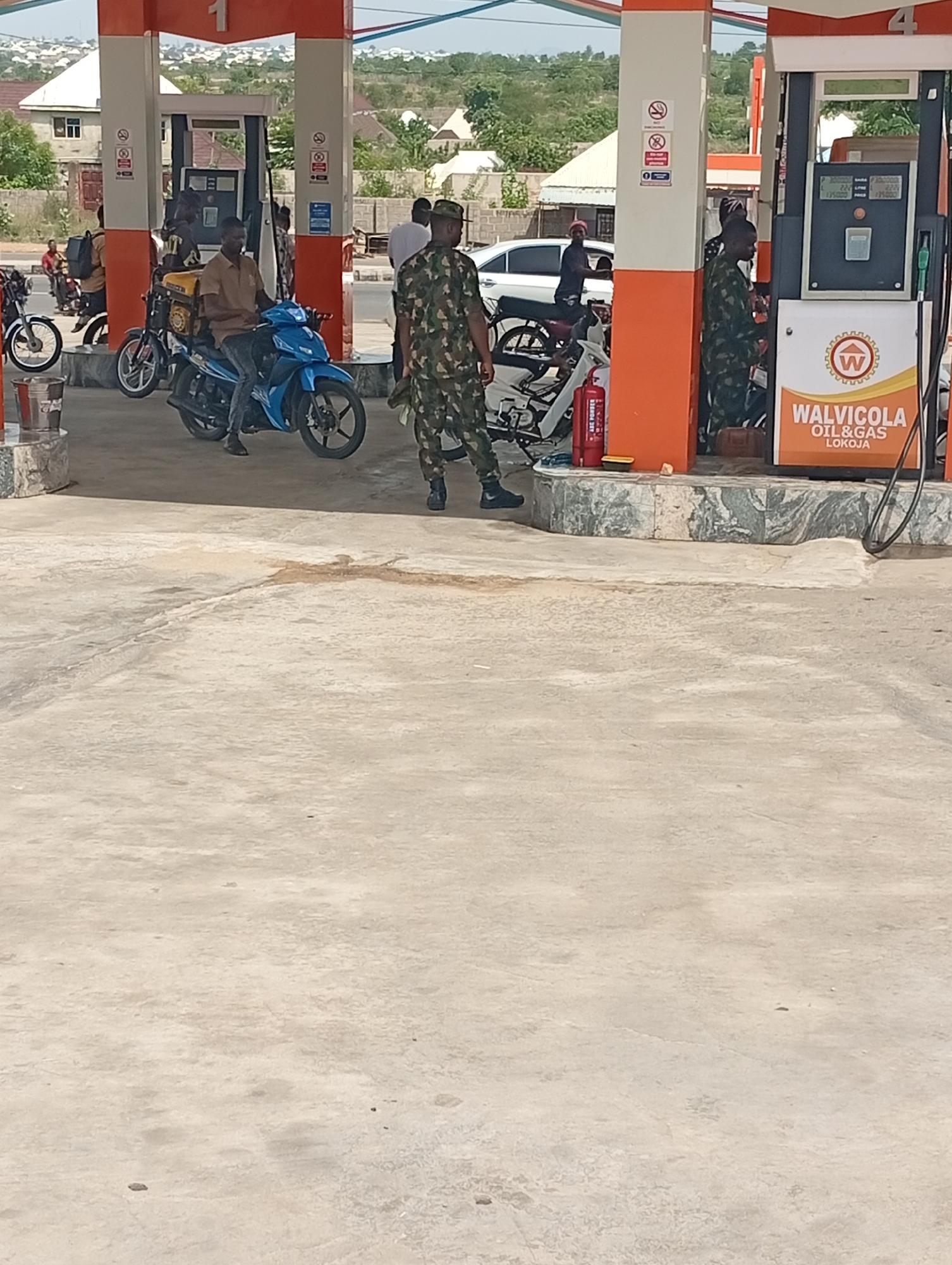 The image shows a gas station in Adankolo, Nigeria, with two fuel pumps under orange and white canopies. A man in camouflage military fatigues is standing near the rightmost fuel pump. A red fire extinguisher is situated between the pumps. In the background, a white car is parked, and several motorcycles are present. One man is seated on a blue motorcycle next to the left pump. There are buildings and trees in the background. The ground is concrete.