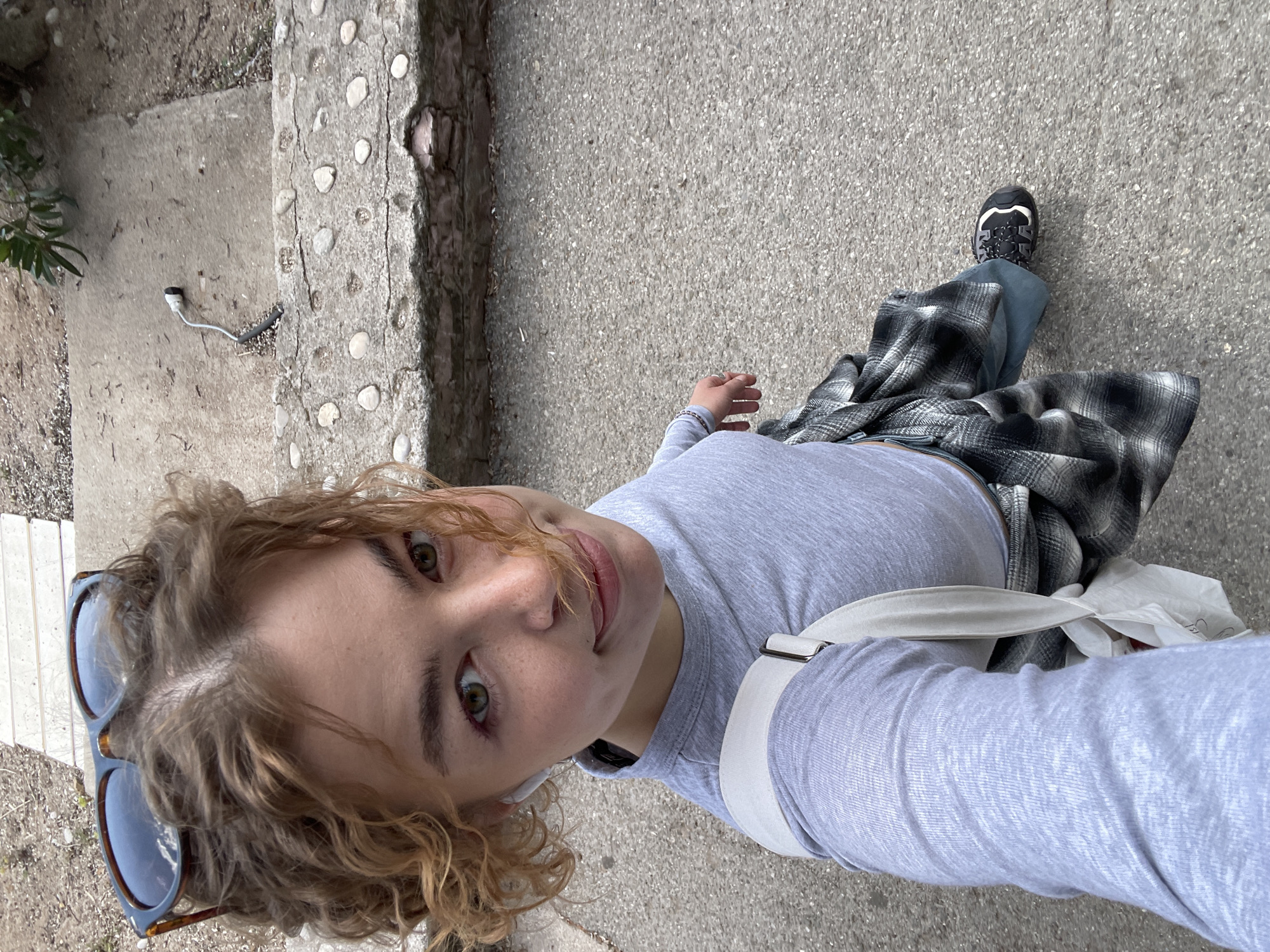 This is a high-angle selfie captured outdoors in Budva, Montenegro, showing a young person looking directly at the camera with a slight smile. The subject, likely female, has light brown to blonde curly hair, with sunglasses resting on top of her head. She has prominent green eyes and is wearing a light grey long-sleeved t-shirt, blue jeans, and a black and white plaid shirt tied around her waist. Black and white athletic shoes are visible on her right foot, and she carries a white shoulder bag with a strap visible across her chest. A white wireless earbud or charging cable is partially visible near her left ear.
The setting consists of a textured grey concrete or asphalt ground, which features small embedded pebbles. To the left, there's a distinct border or path made of rough concrete embedded with numerous rounded white stones, resembling decorative cobbles. A small plant with green leaves and some dry foliage are visible in the upper left background.
The image appears to be taken during the daytime, under bright, natural light, suggesting clear weather. The person's expression conveys a calm and slightly cheerful mood. A white cable with a black tip lies on the ground near the edge of the patterned concrete path. No visible text is present in the image.
