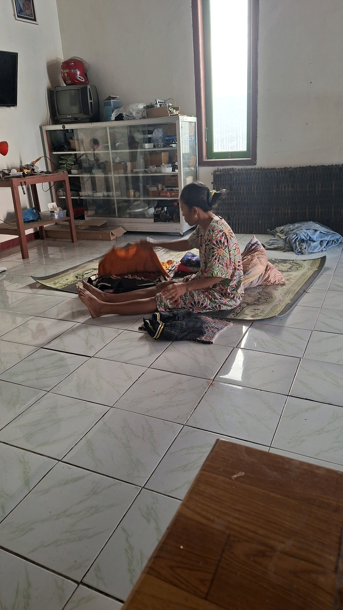 In Sambeng, Indonesia, a woman sits cross-legged on a mat on a tiled floor. She is wearing a floral-patterned dress and is in the process of folding clothes. In front of her are several items of clothing, including an orange shirt and a pair of dark pants. Behind her, a glass-fronted cabinet contains various items, and a vintage television is visible. The room appears to be a living space, with a window letting in natural light. A wooden table is partially visible in the foreground.