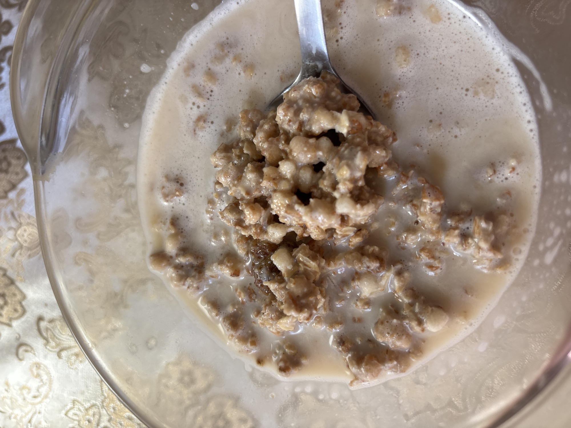 A close-up, overhead shot shows a clear glass bowl filled with oatmeal and milk. A silver spoon is partially submerged in the oatmeal, with a spoonful of the mixture lifted slightly above the bowl. The oatmeal appears to be made with coarse grains, and a few darker pieces, possibly raisins or nuts, are visible. The milk is frothy around the edges of the bowl and the oatmeal. The bowl rests on a tablecloth with an ornate, textured pattern in beige and gold. The lighting is soft and natural, highlighting the creamy texture of the oatmeal and the sheen of the glass bowl. There is no indication of a specific location in Russia.