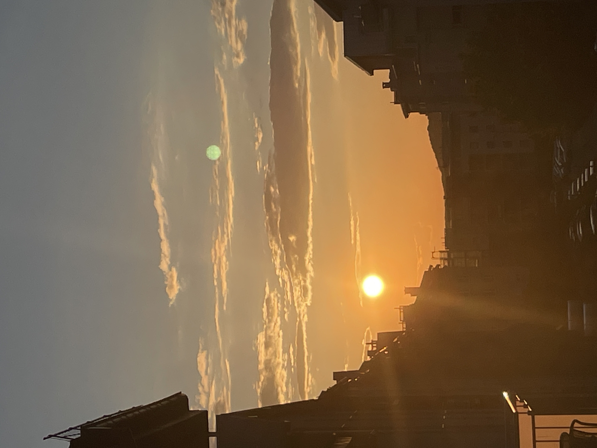 A vertical-oriented medium shot captures a golden sunset scene in an urban environment. The sun is positioned in the center-right, radiating a bright light that permeates the sky. Silhouetted buildings frame the right side and bottom of the image. Wispy clouds stretch across the sky from the top to the bottom. There is a green orb in the upper-left quadrant of the scene. The precise location within the city is indeterminable based on the provided visual data. No individuals or specific actions are detectable within this scene.