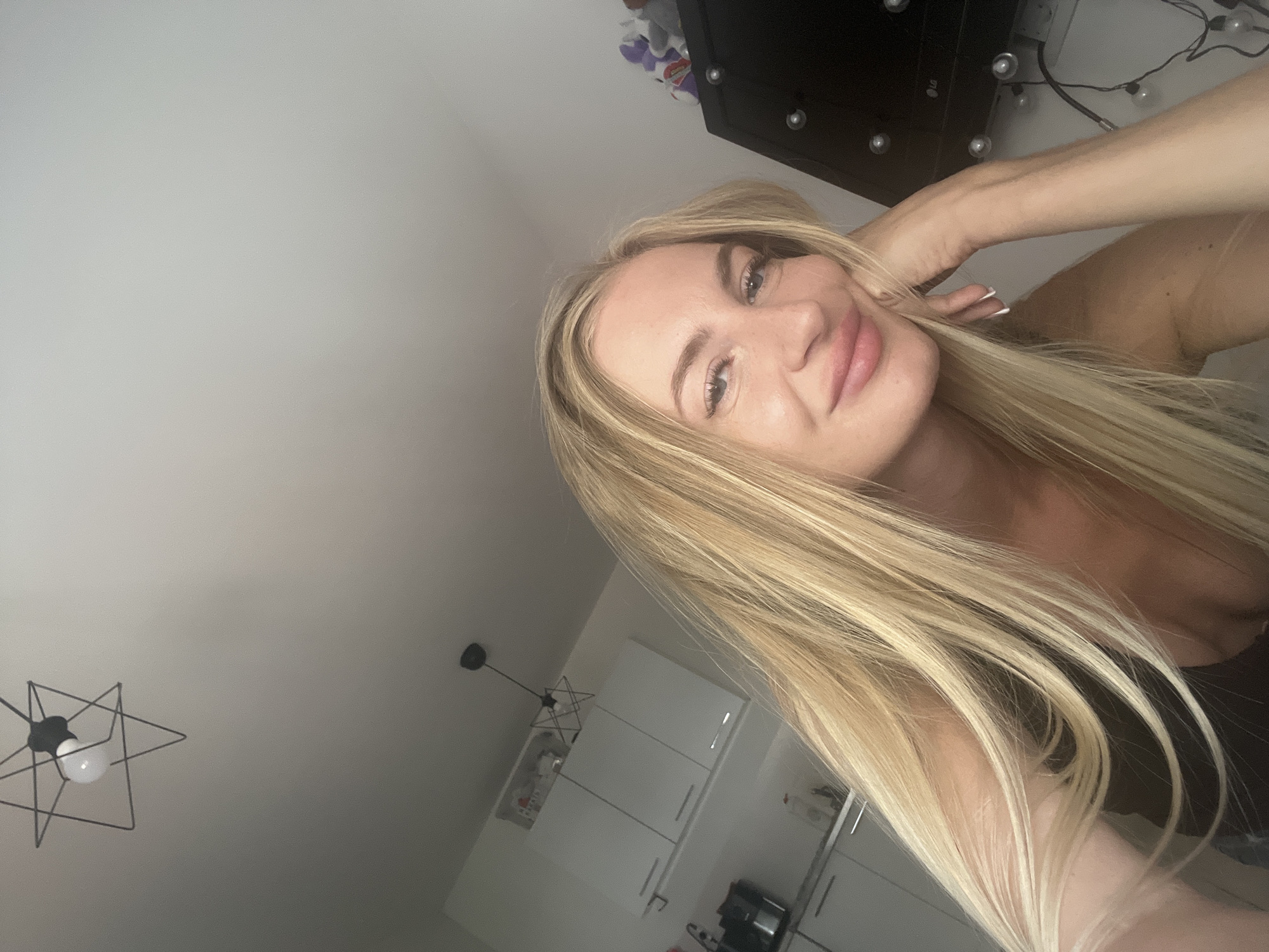 A woman with long blonde hair rests her chin on her hand, looking at the camera. She appears to be indoors, with a kitchen area visible in the background. The kitchen features white cabinets and countertops, along with a coffee maker and bottles on a shelf. A modern pendant light fixture hangs from the ceiling. A black television screen with lights around it is also visible.