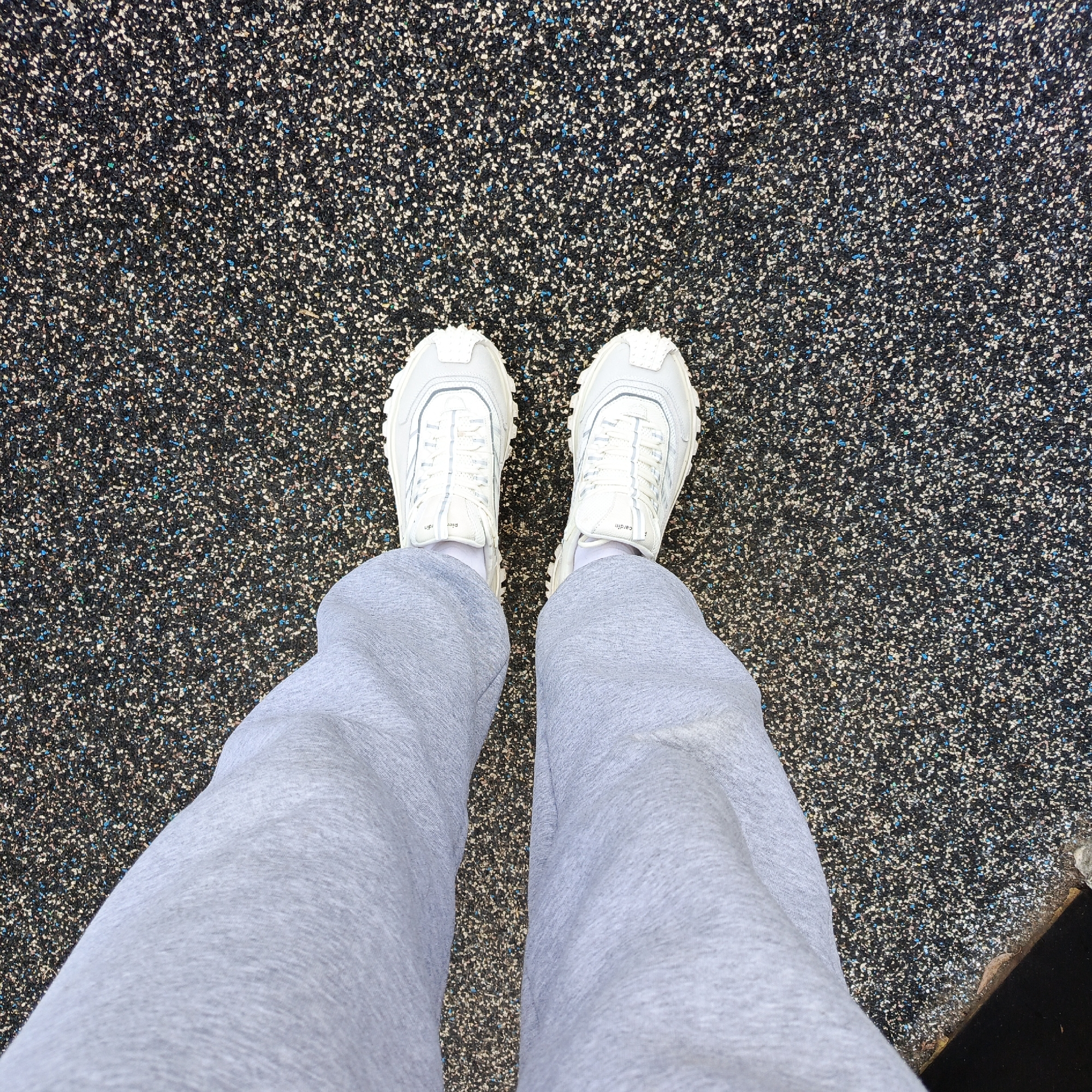 A top-down forensic view depicts the lower extremities of a person standing on a black, granulated rubberized surface in Марковский, Russia. The surface is speckled with white, blue, and minor green particles. The individual is wearing light grey sweatpants or track pants, white ankle socks, and white athletic sneakers with prominent textured soles. The visible branding on the right shoe's tongue includes "Pierre Cardin". A dark, angular object or edge is partially visible in the bottom right corner of the frame.