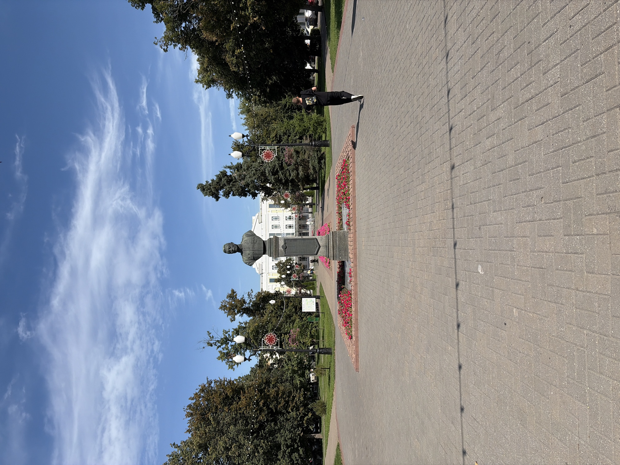 A bust of a man stands on a pedestal in the center of a paved plaza, surrounded by flowerbeds. In the background, a large white building with yellow accents is visible, flanked by trees and lampposts. A person wearing a black t-shirt and pants is jogging away from the camera on the right side of the plaza. The sky is blue with scattered white clouds. This is likely a public space in the city of Tambov, Russia, given the context of the uploaded image and common architectural styles in Russian cities.