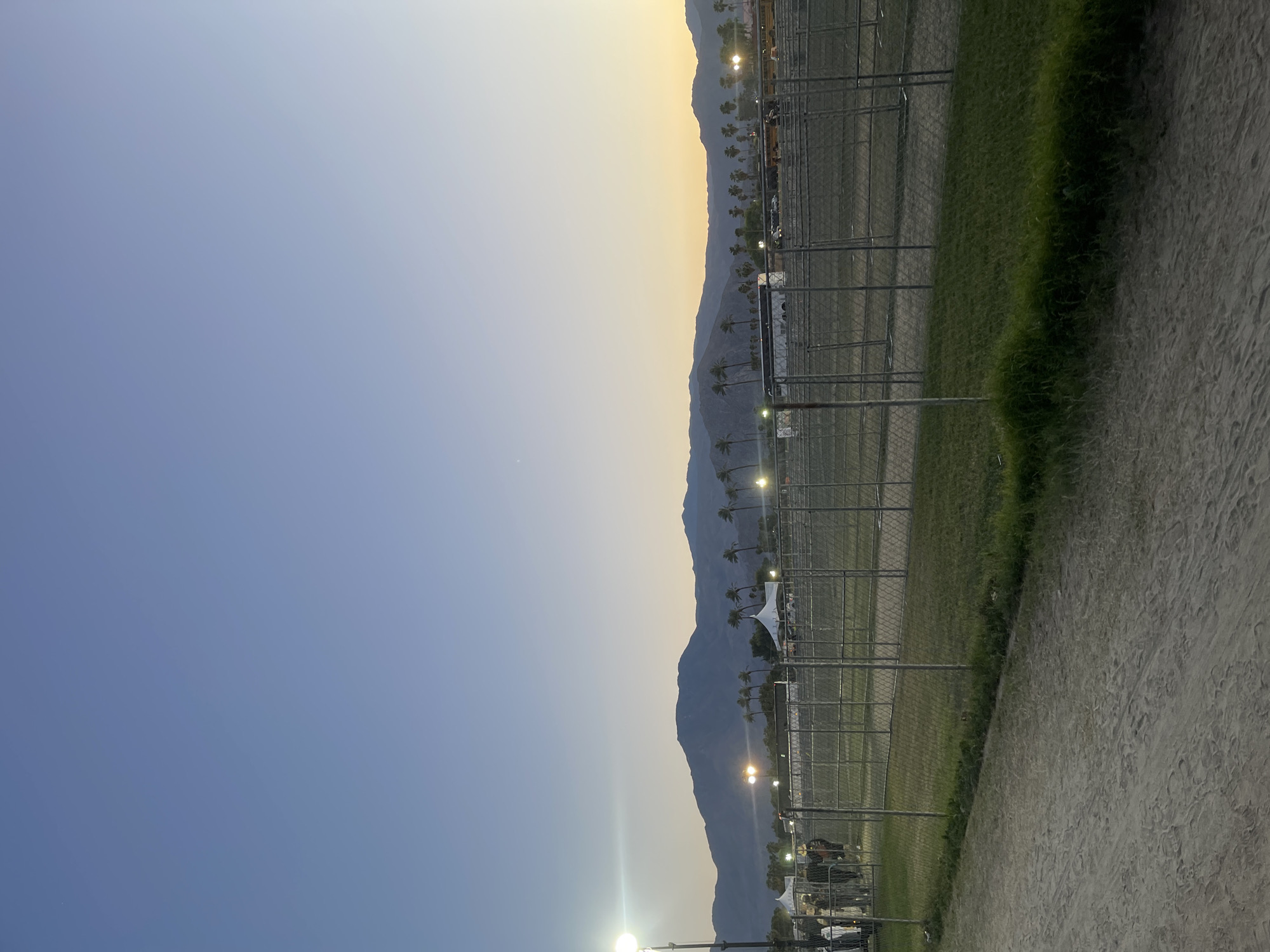 The photograph captures a twilight scene in Indio, California, United States of America. Mountains are visible in the background, their forms outlined against a gradient sky transitioning from a golden hue near the horizon to a blue above. A chain-link fence runs along the right side of the frame, separating a grassy area from a sandy or dirt path. Scattered palm trees are interspersed throughout the area. Artificial lights punctuate the landscape, suggesting activity in the area.