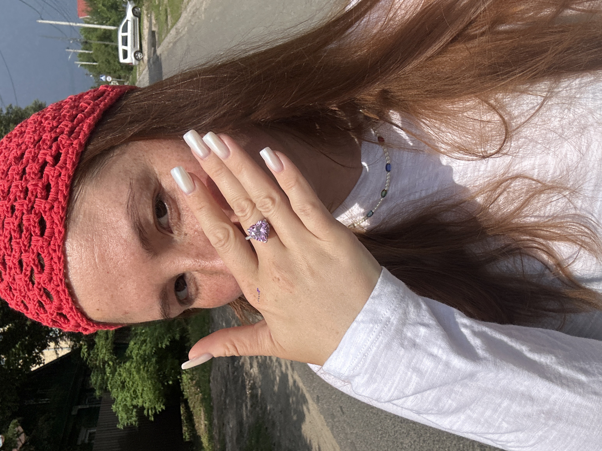 A woman with long, auburn hair, wearing a red crocheted beanie and a white long-sleeved shirt, is pictured outdoors. She is partially covering her face with her left hand, revealing her eye, nose, and forehead which have freckles. On her left hand, she has long, square-shaped, pearlescent white nails and is wearing a ring with a heart-shaped purple stone. A pearl necklace with multi-colored beads is visible around her neck. The background shows a street with a paved surface and a white minivan in the distance. Trees and a fence are also visible on the left side of the image, and a dark, cloudy sky is present in the upper right corner. The environment suggests a sunny day with a storm approaching. The specific location in Nizhny Novgorod, Russia cannot be determined from the visual information provided.