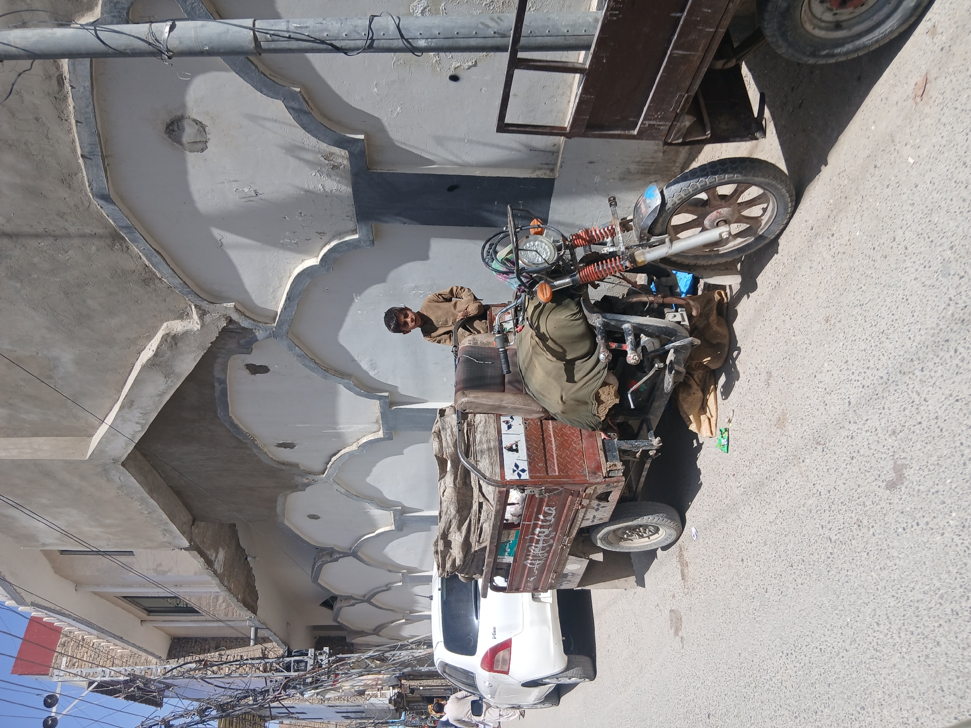 A three-wheeled motorcycle is parked on a street next to a white car in Quetta City Tehsil, Pakistan. The vehicle is heavily loaded with items covered by a tarp. A man is sitting on the motorcycle, looking to the left. Overhead are tangled electrical wires and the eaves of a building, which are decorated with an alternating pattern of smooth white curves and darker gray recessed areas. The street is light gray and appears to be asphalt.