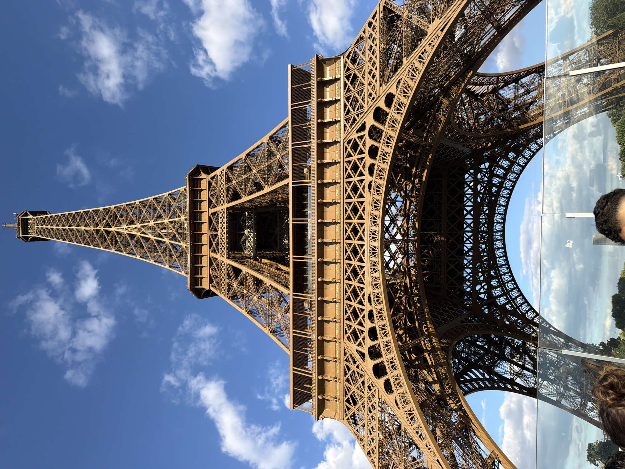 This media file shows a low-angle, upward view of the Eiffel Tower in Paris, France. The iconic iron lattice structure dominates the frame, set against a clear blue sky dotted with white cumulus clouds. The sun is shining, indicating it is daytime, likely late morning or early afternoon given the bright light and clear sky. The architecture of the tower is detailed, with its intricate metalwork clearly visible.
The reflection in a glass surface on the right side of the frame offers a mirrored view of the tower and the surrounding parkland, including trees and sky. A person's head and shoulders are partially visible in the bottom right corner, suggesting the image was taken by a tourist or visitor to the landmark. The overall mood is one of awe and appreciation for the historical monument. The scene is peaceful and scenic, capturing a classic Parisian vista. No visible text is present in the media file.