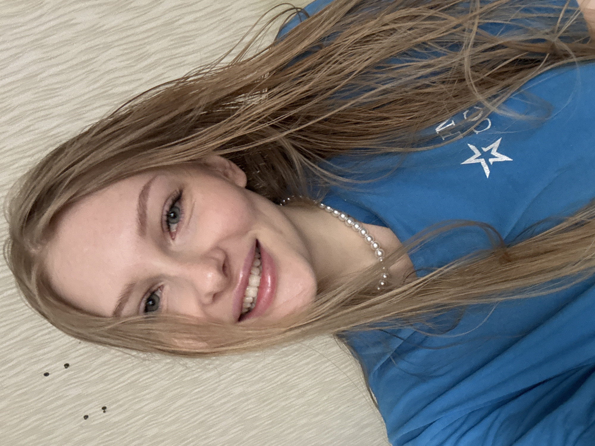 A young woman with long, light brown hair is pictured lying down, smiling broadly at the camera. Her head is tilted, and her blue-green eyes convey a cheerful emotion, complementing her wide smile which reveals white, even teeth. She appears to be wearing minimal makeup.
She is dressed in a vibrant blue top or t-shirt, and a simple string of small, round pearls adorns her neck. On the blue fabric of her top, partially covered by her hair, white letters are visible along with a distinct white five-pointed star logo. The visible letters appear to be "CO", suggesting part of a brand or word.
Her long, light brown hair is somewhat tousled and spread out around her head, appearing natural and slightly damp.
The background consists of a light-colored, possibly cream or off-white, surface with a subtle, wavy texture, likely a blanket or bedspread. A few small, dark specks are scattered on this surface near her head. The image depicts an indoor, casual setting, located in Кондратово, Russia.