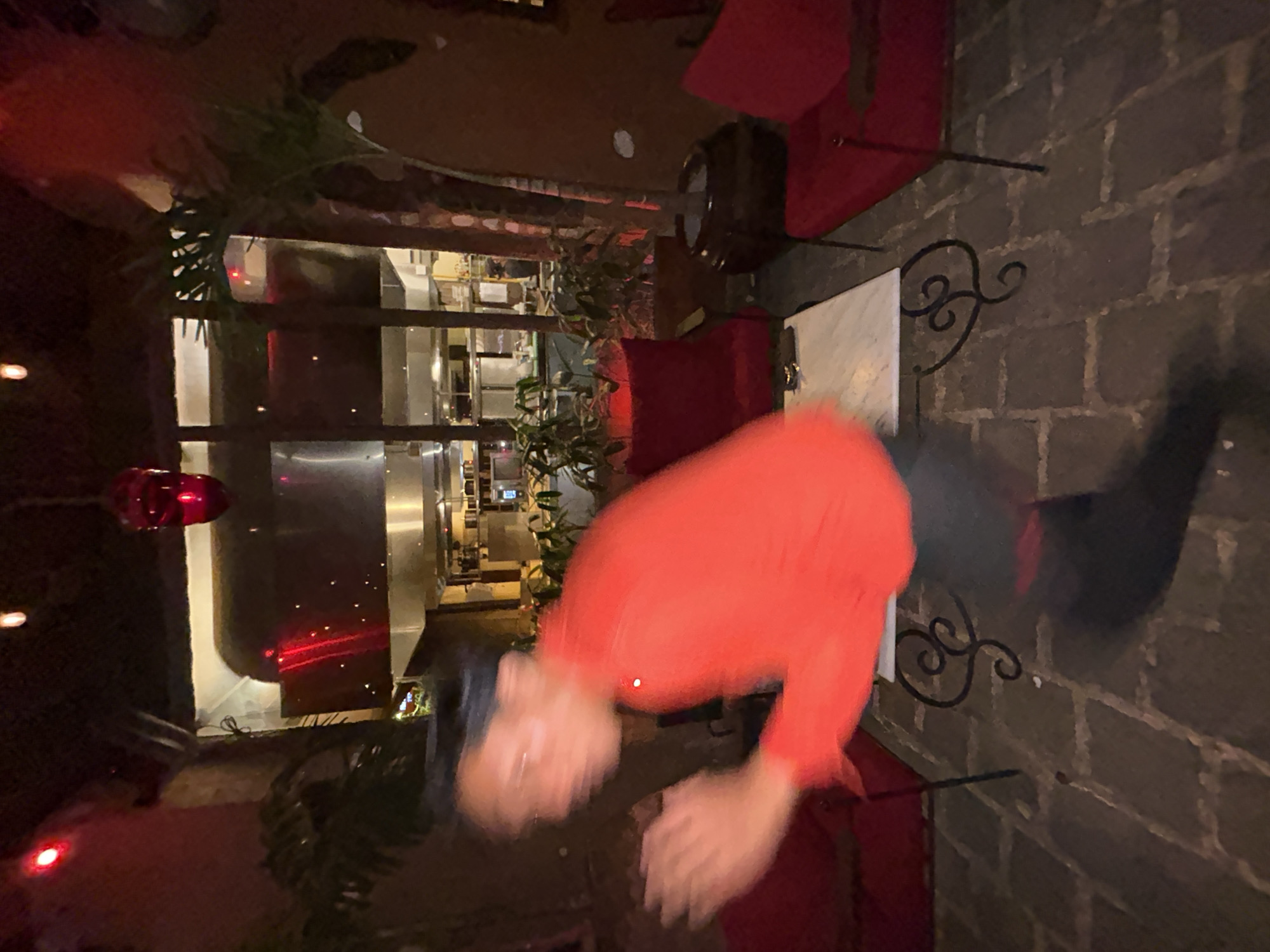 The image depicts an outdoor dining area at night, with a blurred figure in the foreground wearing a red shirt and dark pants, appearing to be in motion. Behind the figure, a kitchen area is visible through a glass partition. The environment includes red chairs, potted plants, and a marble-topped table. The flooring is paved with rectangular stones. The scene suggests a restaurant setting in Mengwi, Indonesia, characterized by low lighting and a focus on the dining and kitchen areas.