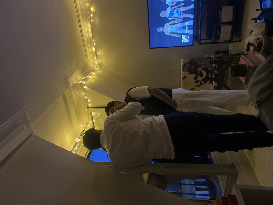 Two men stand in a room adorned with string lights. One wears a white shirt and dark pants, gazing out a window. The other sports a white shirt, vest, and white pants. A television screen displays images of people, potentially a sports team wearing blue uniforms with "Waterloo" visible. The room's decor includes a cowhide rug, a small table with a plant, and various stacked boxes.