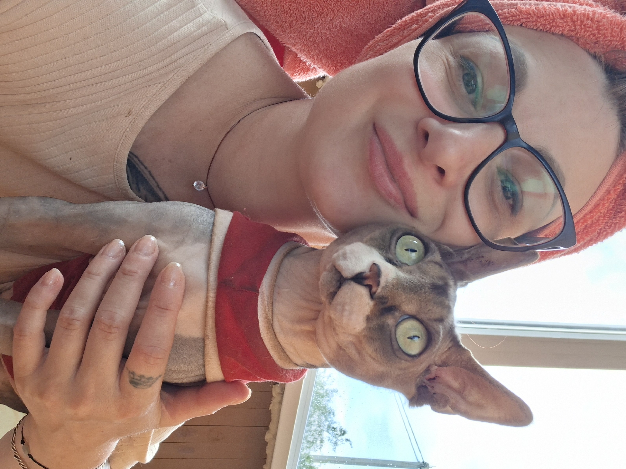 In a close-up, outdoor portrait, a woman with a bright pink towel wrapped around her head, wearing black-rimmed glasses and a light beige ribbed tank top, smiles at the camera while holding a hairless cat. The cat, a Sphynx, has large, round, light green eyes and is wearing a red and white garment around its neck and chest. The woman has a thin necklace with a clear pendant. Behind them, a window shows a bright sky with some faint foliage and a wooden wall. A subtle tattoo is visible on the woman's forearm. The image is taken from a low angle, focusing on the subjects.
