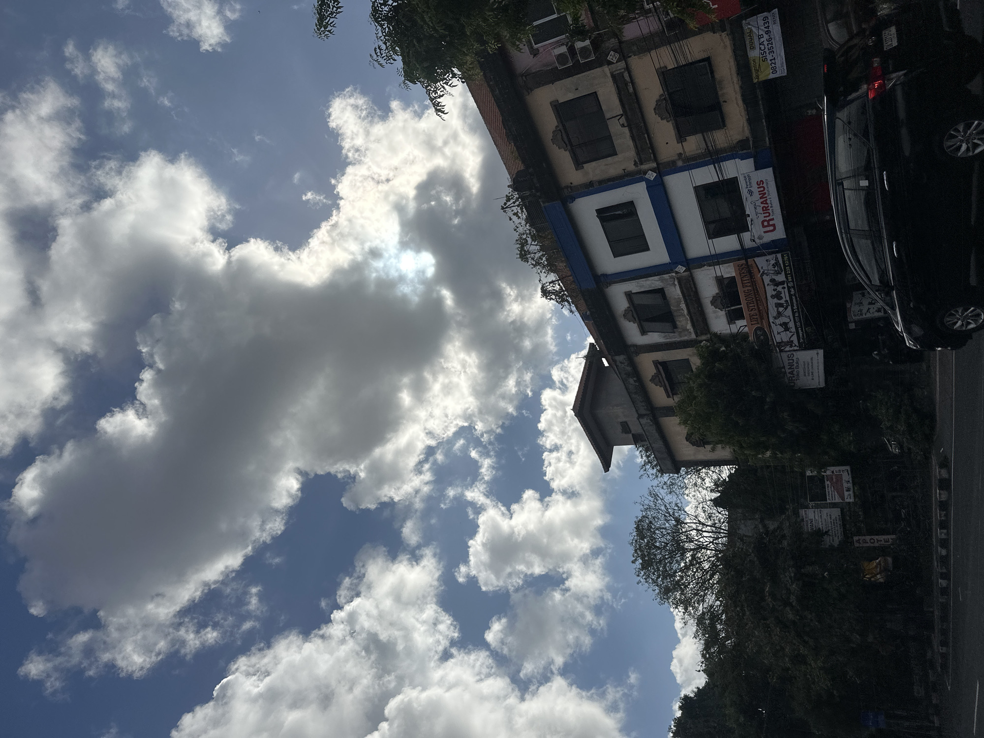 This image captures a street scene in Denpasar, Indonesia, with a focus on a row of multi-story buildings against a backdrop of a partly cloudy sky. The buildings appear to house various businesses, with signage visible, including "LIFE STRONG FITNESS" and "URANUS" which is labeled as "Toko Buku Stationery" (Bookstore Stationery). One building has a blue and white facade, while others are painted in earthy tones. Trees and foliage are present, partially obscuring some of the structures. A black car is parked or driving on the road in the foreground, and the street appears to be paved with a white and black checkered curb. The sky is a mix of bright blue and dramatic, large white and gray clouds, with the sun peeking through in one area, creating a bright spot. The composition is angled, giving a dynamic perspective of the urban environment and the sky above.