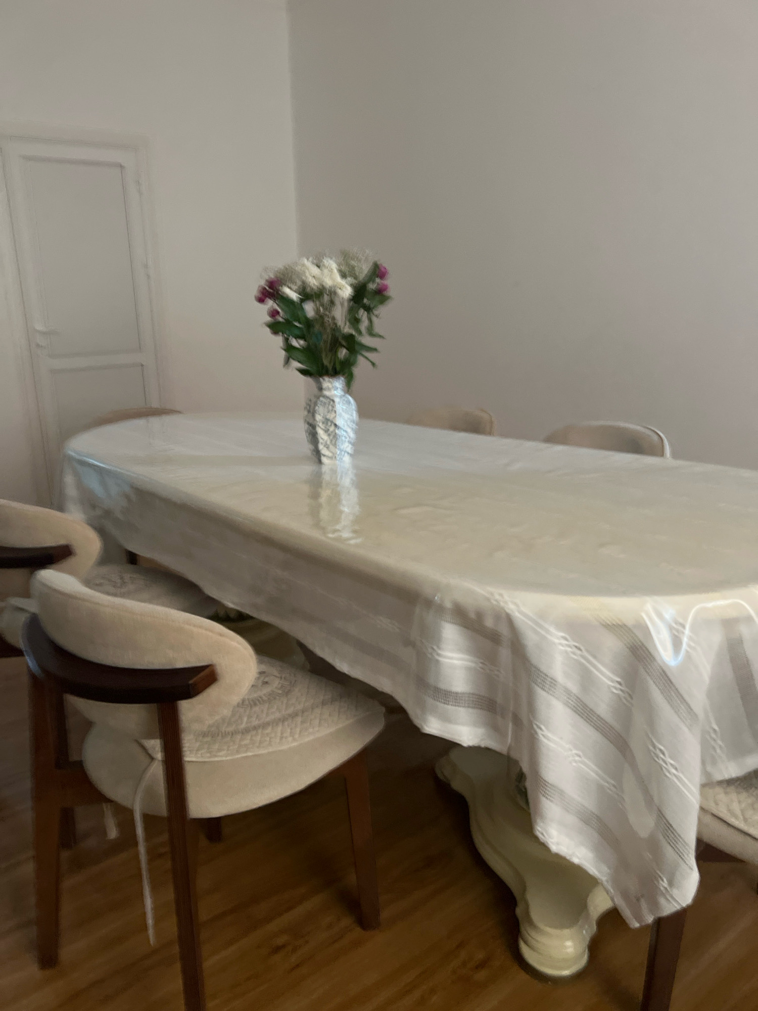 An oval dining table draped with a clear plastic tablecloth and a white decorative overlay is set in a room with pale walls and a wooden floor. A patterned vase holding a bouquet of white and pink flowers sits centrally on the table. Several chairs with upholstered seats and dark wooden frames surround the table. In the background, a white door is visible on the left. The scene is dimly lit, suggesting an indoor setting during daytime or evening. There is no indication of the city of Novogagatli, Russia.