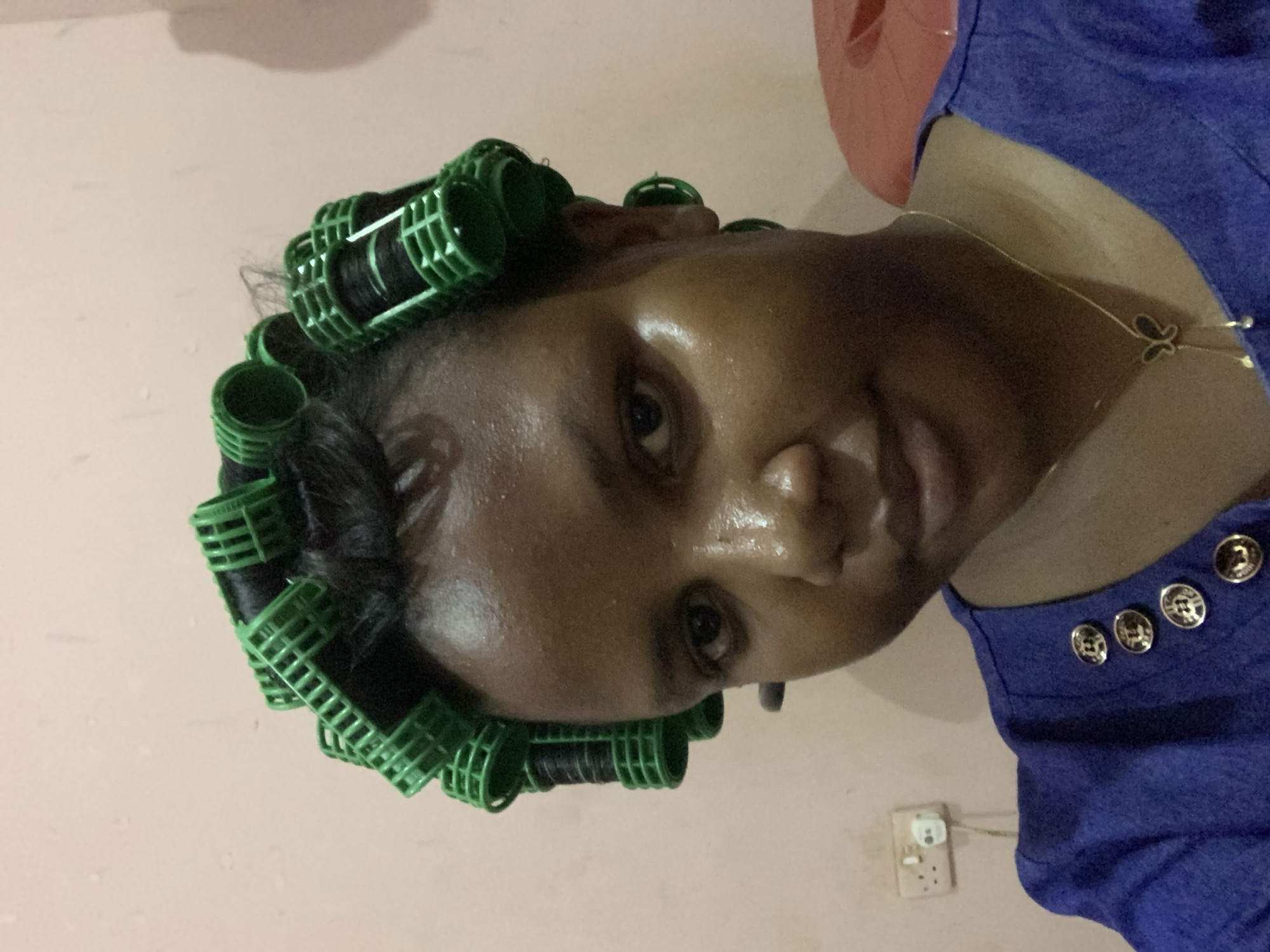 A close-up, slightly angled portrait of a dark-skinned woman wearing green hair rollers. Her hair is black, wrapped around the rollers. She is wearing a blue shirt with silver buttons on the shoulder. A delicate gold necklace with a butterfly pendant is visible. The background is a peach-colored wall with an electrical outlet. The location appears to be indoors. There are no indications of being in Dar es-Salaam, Tanzania.