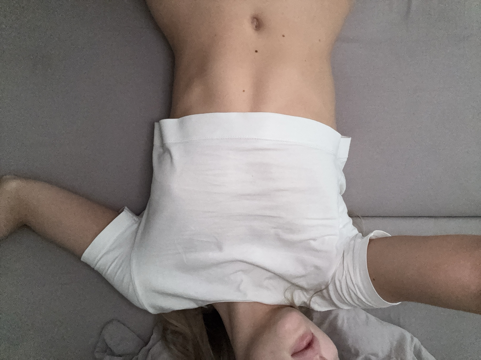 The image is an upside-down, close-up, eye-level shot of a person lying on a gray sofa. The person is wearing white underwear and a white t-shirt. The person's bare torso, midriff, and legs are visible. They are lying with their legs spread apart. The person's face is partially visible at the bottom of the frame, turned to the side. The lighting appears to be natural, suggesting it could be daytime. There is no visible text in the image. The setting is likely a home due to the presence of a sofa and the casual attire of the person.