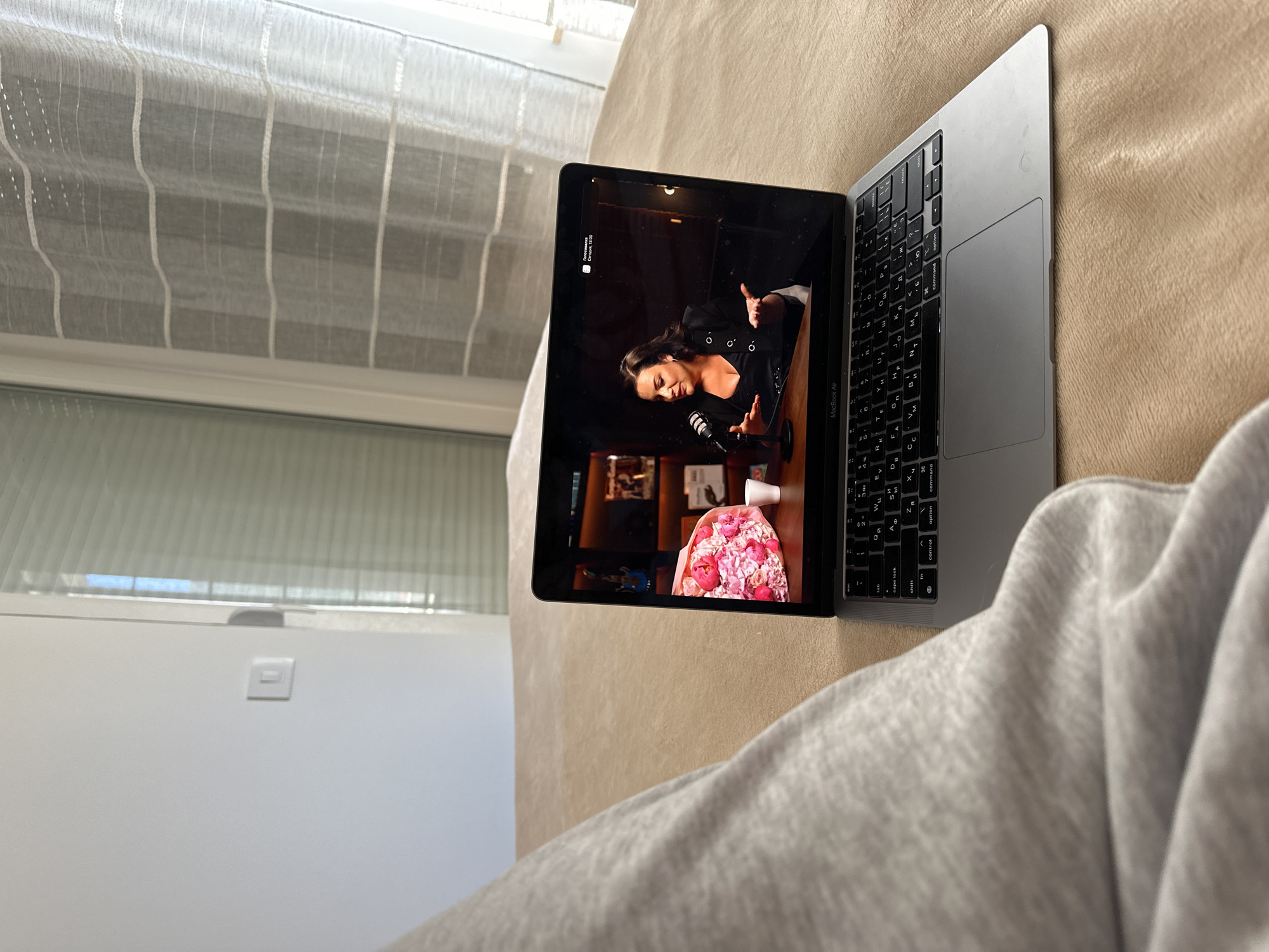 A laptop displays a video of a woman in a black jacket speaking into a microphone. The laptop sits on a tan sofa. A person wearing a gray sweatshirt is visible in the foreground. A window with a partially open white blind is to the left. This scene appears to be inside a home in Donji Pobori, Montenegro, based on the location information.