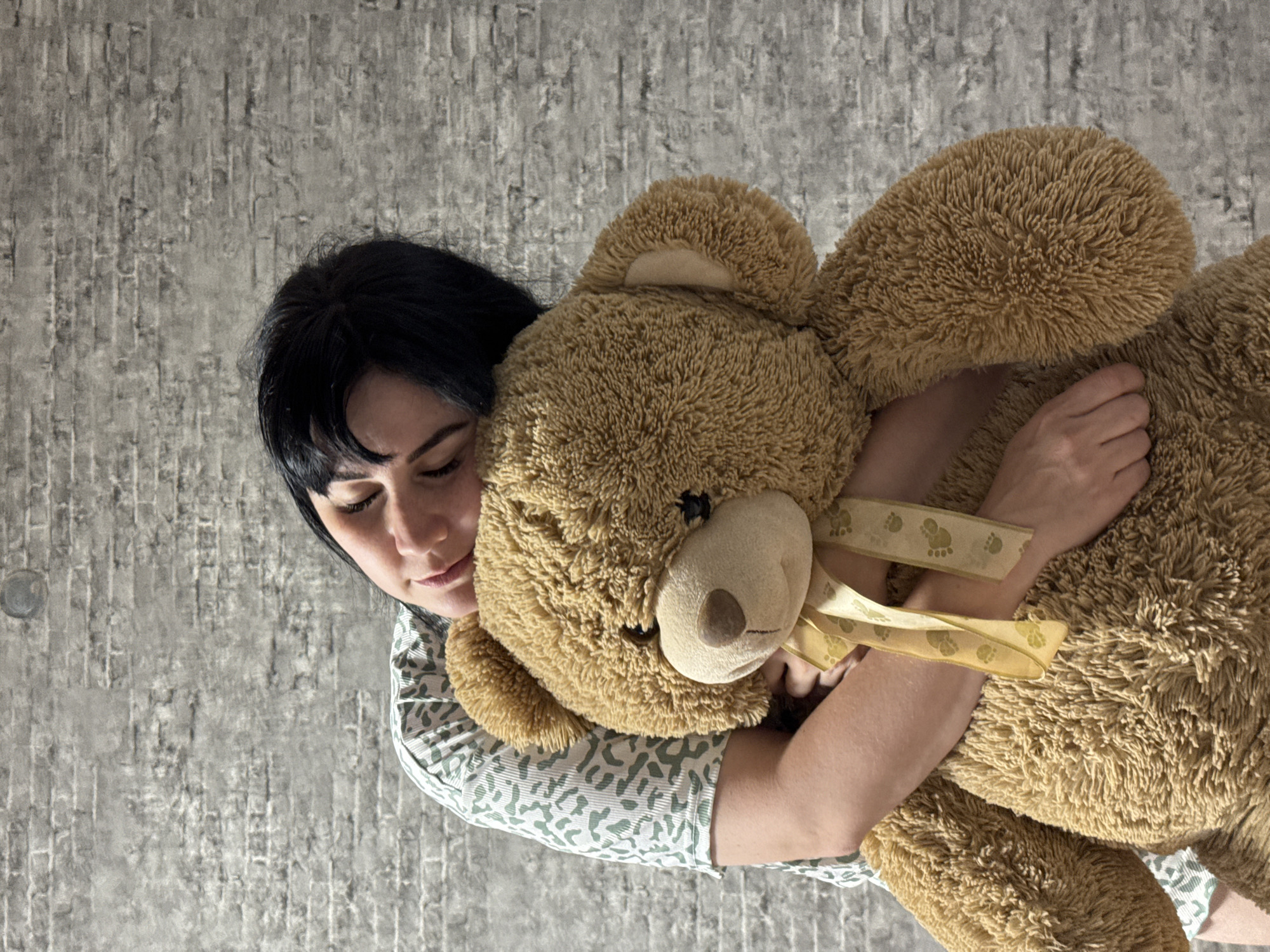 A woman with dark hair is embracing a large brown teddy bear. She is wearing a light-colored shirt with a green pattern. The background is a gray wall with a textured pattern. No information indicates the location is Zav'yalovo, Russia.