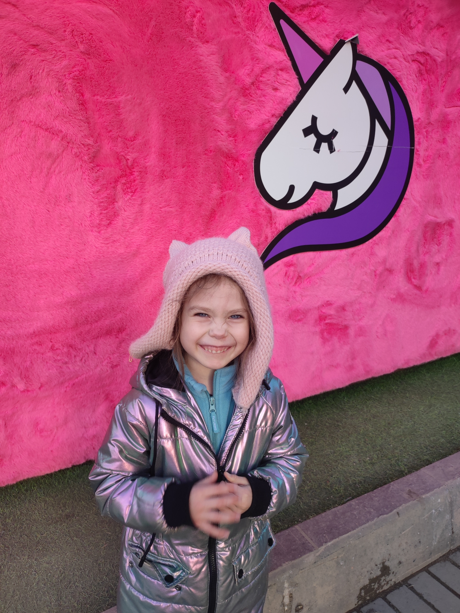 A young girl stands smiling in front of a bright pink display that features a unicorn head, likely in a public area within the city of Votkinsk, Russia. The girl wears a pink knitted hat with ear-like protrusions, a metallic silver winter coat, and a blue zippered jacket underneath. She is holding her hands together in front of her. The background features a large, fuzzy pink surface with a stylized white unicorn head outlined in purple in the upper right corner. The ground appears to be artificial grass adjacent to a concrete ledge or low wall.