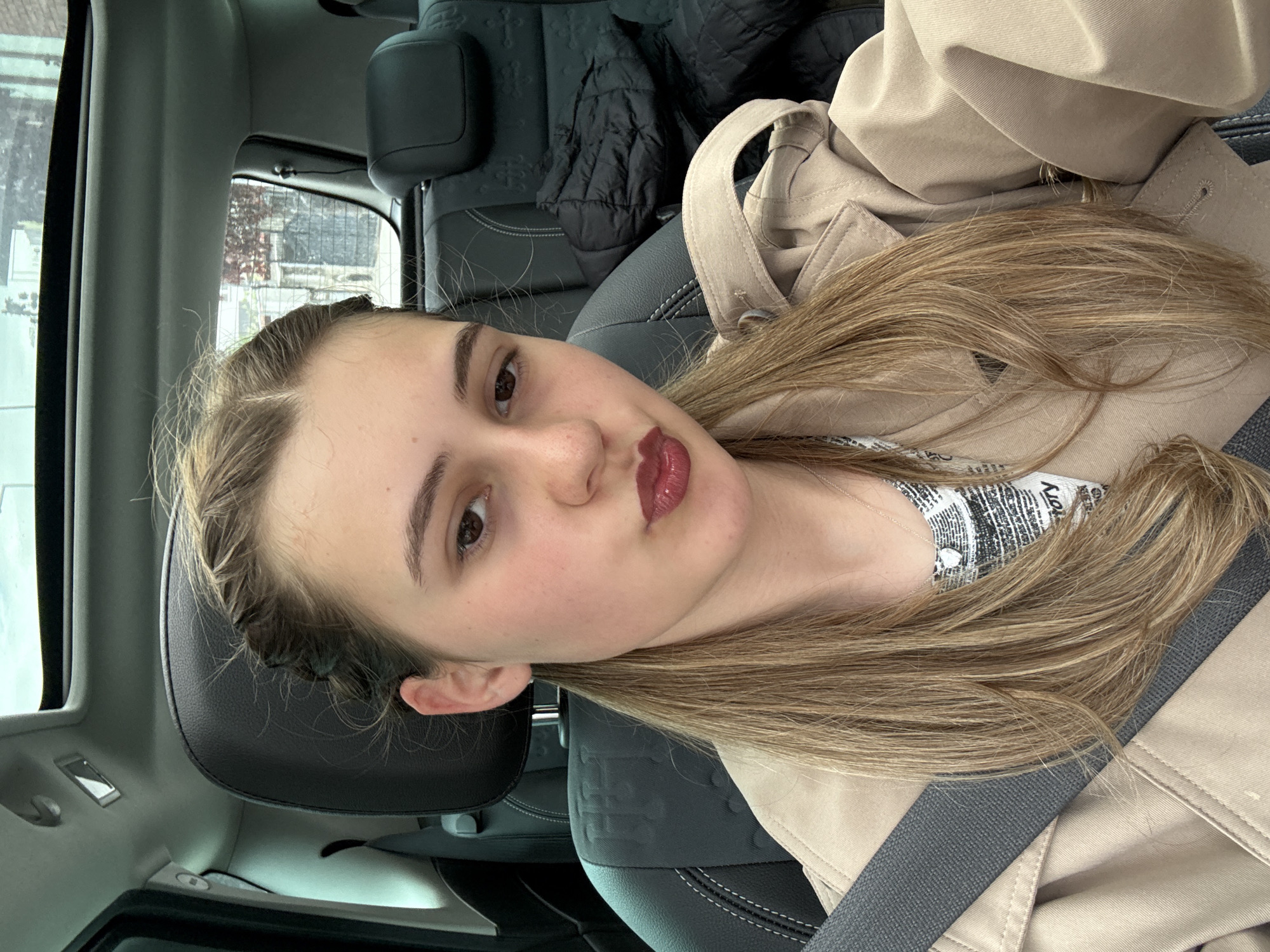 The image is a selfie taken inside a car. A young woman with long blonde hair is sitting in the driver's seat, wearing a beige trench coat and a seatbelt. She has her hair styled with braids close to the face and wears dark red lipstick. A black jacket is visible on the backseat behind the driver's seat. Outside the car window, it appears to be daytime, with a glimpse of buildings and foliage, but no information is given to confirm the location is in Panagyurishte, Bulgaria.