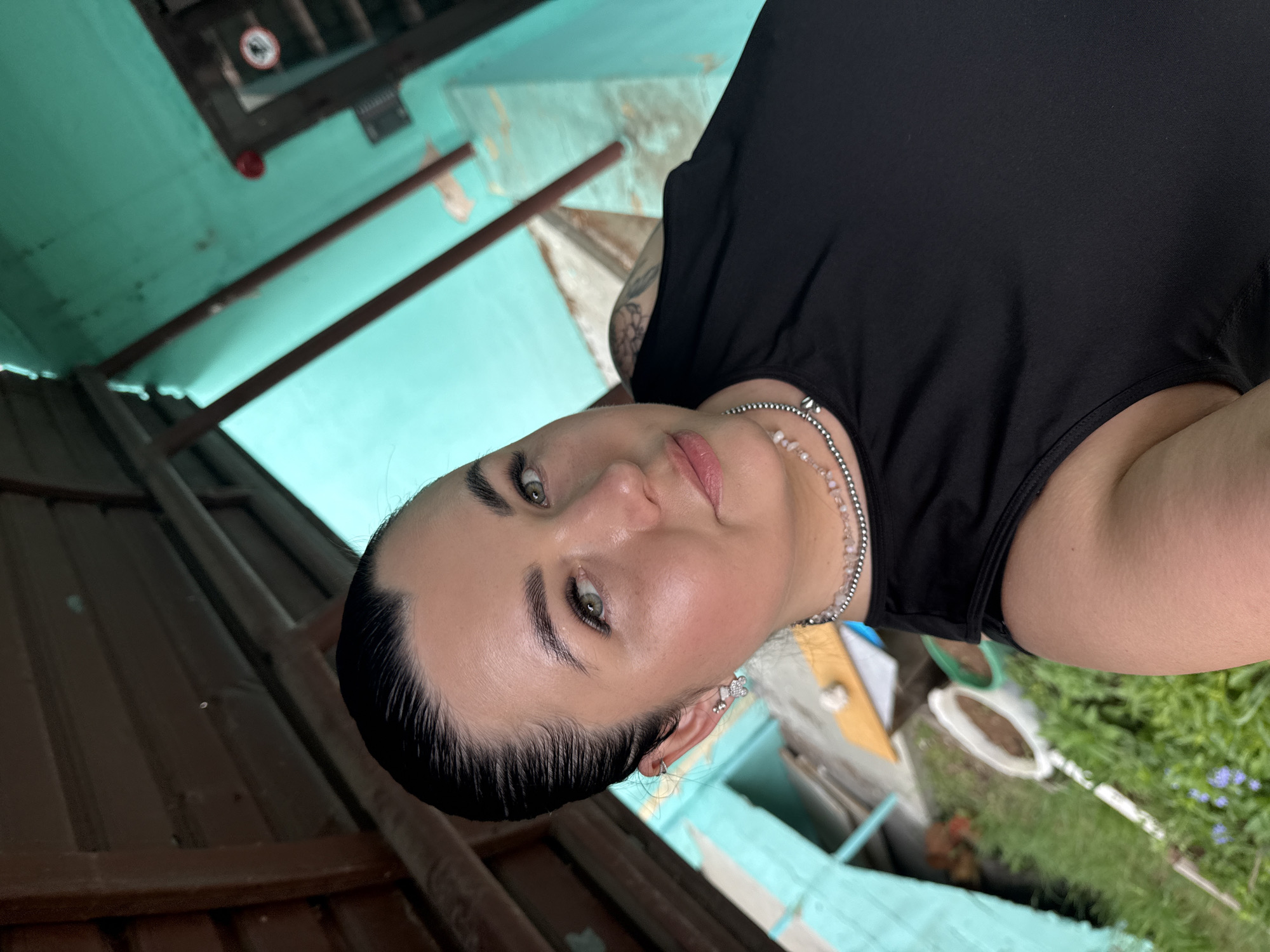 A woman is photographed from a low angle. She wears a black top and a layered necklace. Her hair is dark and pulled back. She has light skin, green eyes, and visible piercings in her ears. In the background, there's a structure with blue walls and brown corrugated metal sheets. A fire safety sign is visible near the ceiling. The setting seems to be outdoors in what could be the city of Chaikovsky, Russia, given the prompt. Greenery is visible in the lower right corner of the image.
