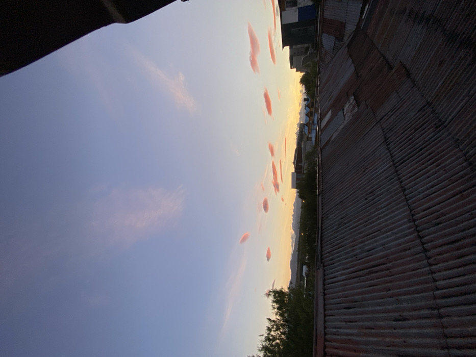 A view of a sunset sky from a rooftop. The sky transitions from deep blue to pale yellow, dotted with pink clouds. A corrugated metal roof dominates the right side of the frame. In the distance, buildings and trees line the horizon. The image is taken from a low angle, with a dark overhang in the top left corner.