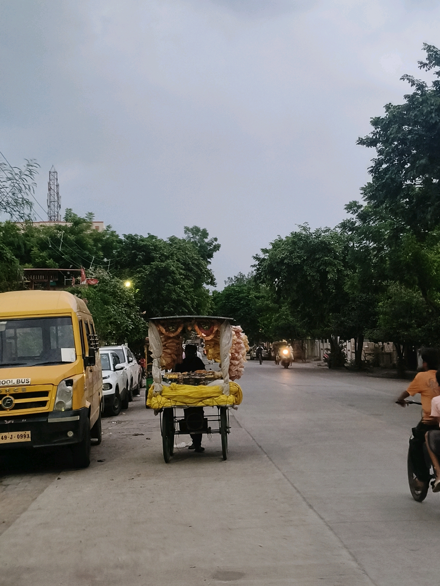 The image depicts a street scene in Nagpur, India, under an overcast sky. In the foreground on the left, a yellow school bus is parked. Next to it, a food cart is visible, pushed by a person wearing a dark shirt. The cart is laden with food items, some wrapped in plastic and others in a yellow covering. The street is paved, and trees line the right side of the road. Further down the street, a person on a motorcycle is visible, and a few other vehicles are parked. The overall ambiance suggests a typical urban environment in India.