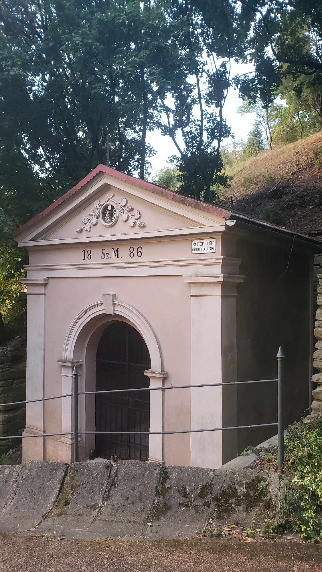 The image shows a light pink, single-story building with a triangular pediment roof. Above the arched entrance, which is covered by a black metal grate, there are inscriptions and decorative elements. To the right of the entrance, a small rectangular sign is affixed to the building. The building is situated on a slight incline, with trees and vegetation surrounding it. A low concrete wall and a metal railing are in the foreground. The building is identified as the "Svidnicka Kaplnka" (Svidnik Chapel) in the city of Prešov, Slovakia.